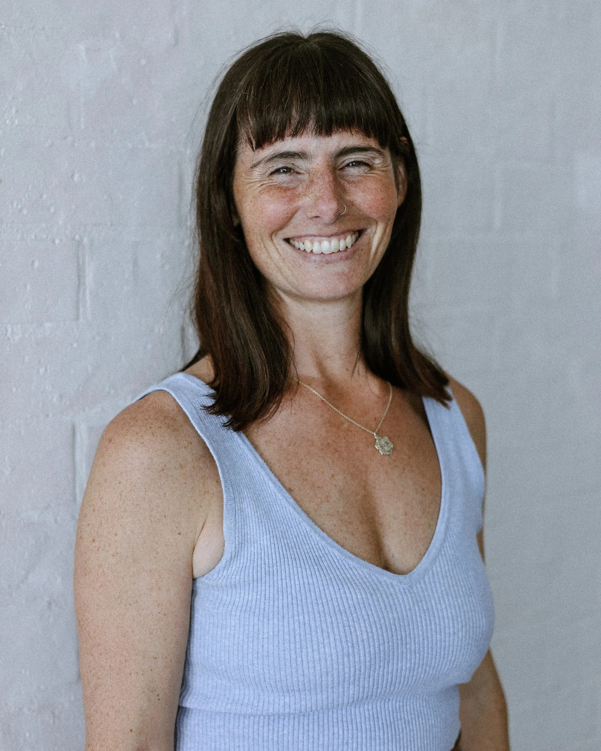 A woman with brown hair and bangs, smiling, wearing a light blue sleeveless top and a gold necklace with a pendant, standing against a plain gray wall.