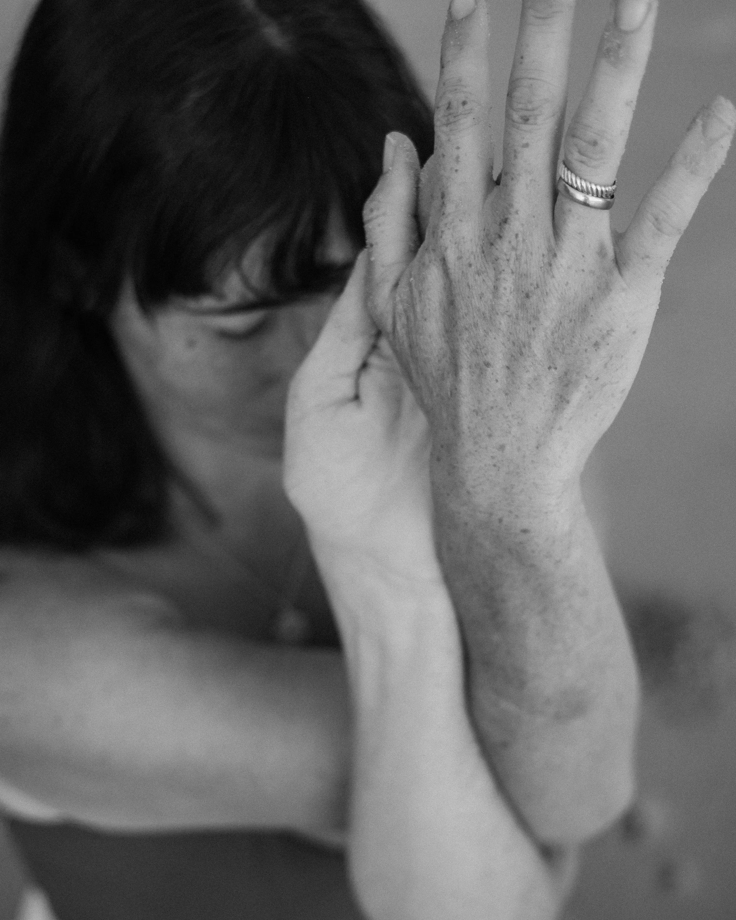 A person with dark hair and glasses resting their head against a large, textured hand that is raised vertically. The hand has a ring on the ring finger, and the image is in black and white.