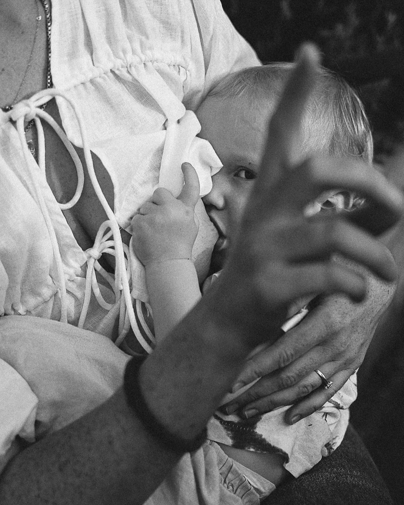 A young child with short hair on a hospital bed, giving a thumbs-up while being breastfed by an woman with a ring on their finger, in a black-and-white photograph. This is a motherhood photography session