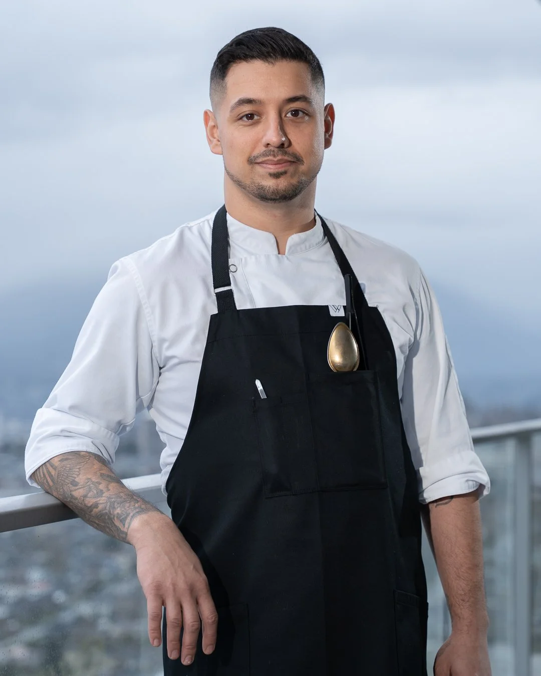 Chef wearing a white chef's coat and black apron standing outdoors with cityscape in the background.