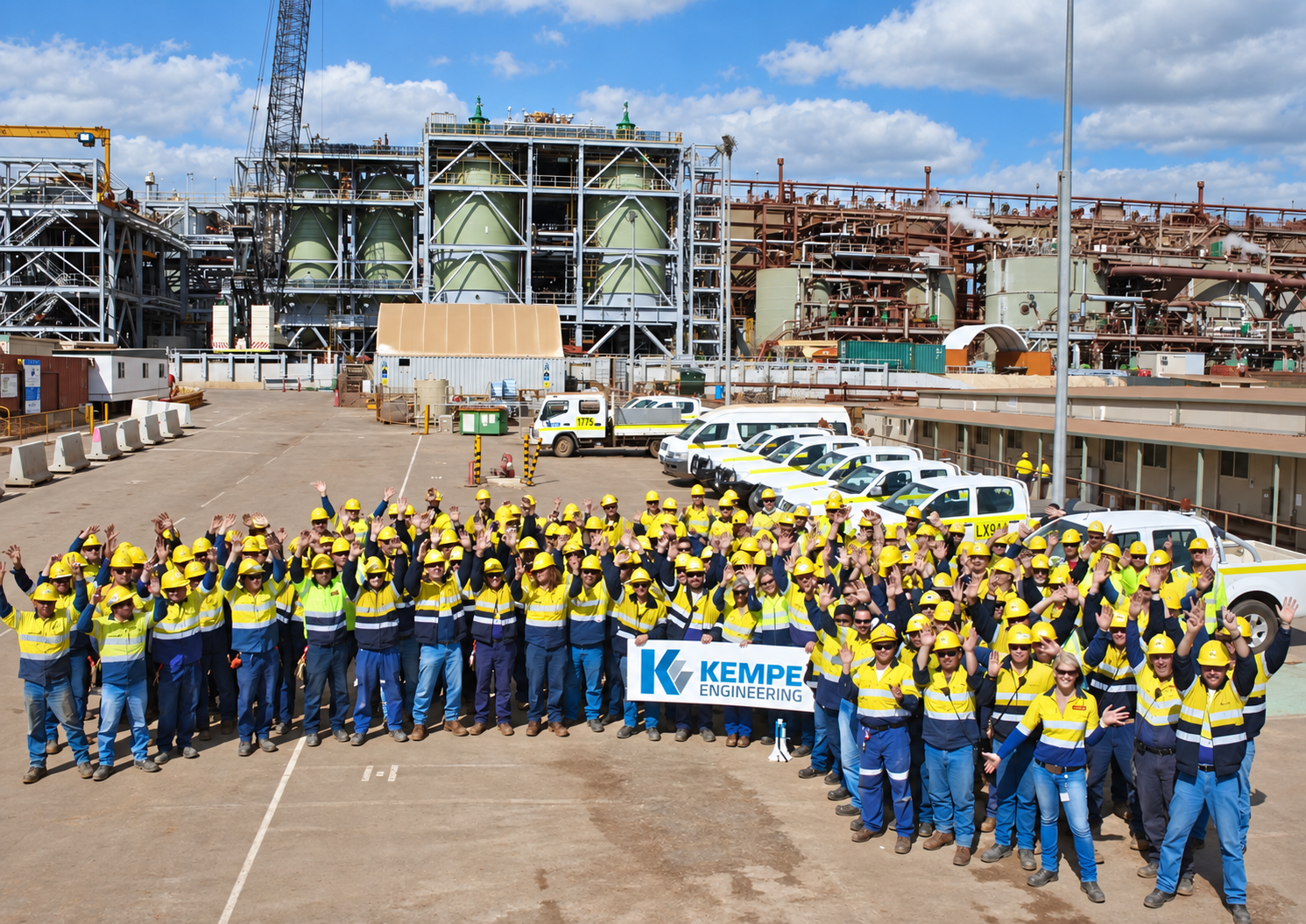 Group of workers in yellow safety helmets and high-visibility vests smiling and waving, holding a "KEMPE ENGINEERING" banner, in front of an industrial plant with machinery, vehicles, and equipment on a bright, partly cloudy day.