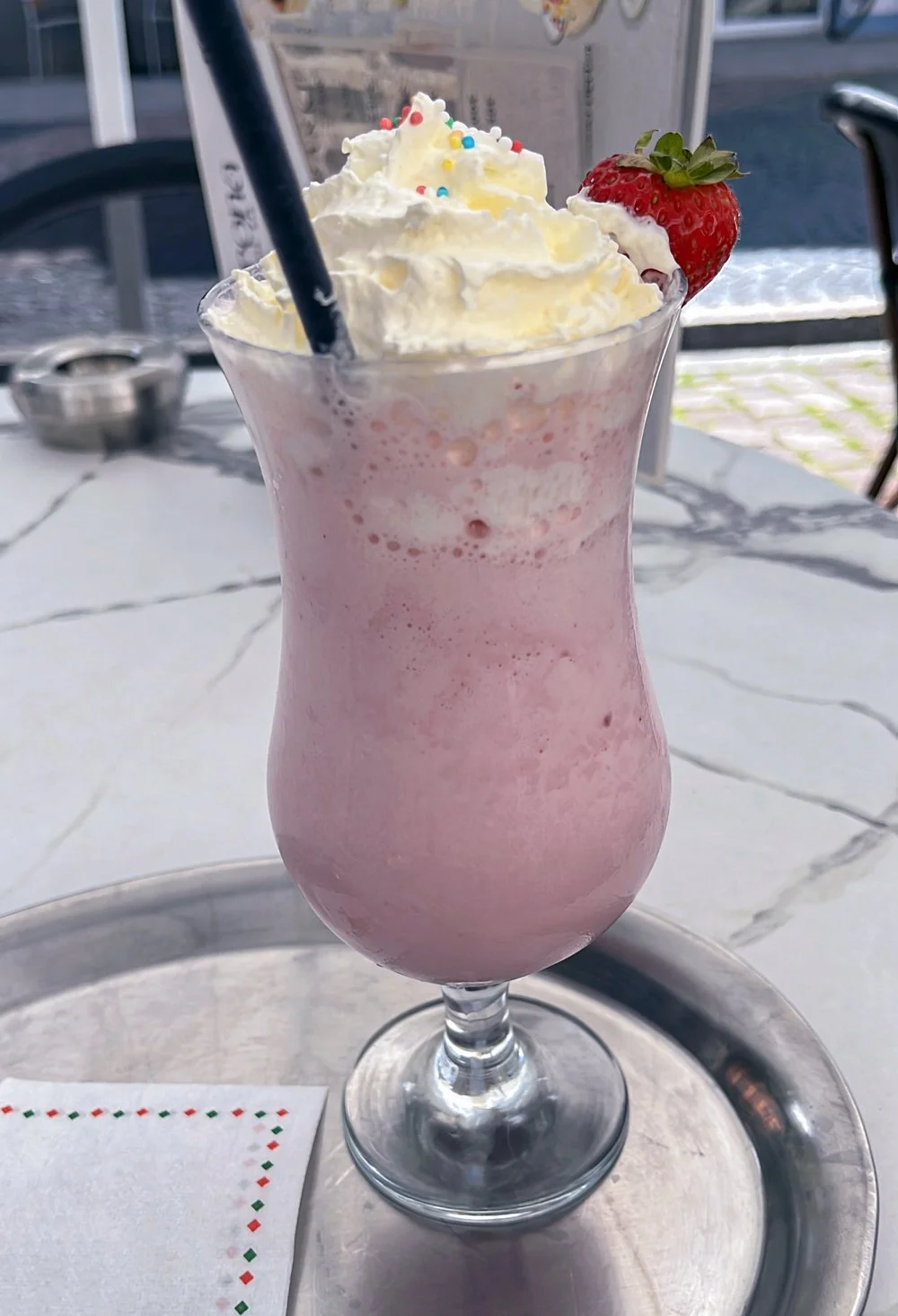 Ein Glas mit Erdbeer-Milchshake, garniert mit Schlagsahne, bunter Streuseln und einer Erdbeere, auf einem Tablett.