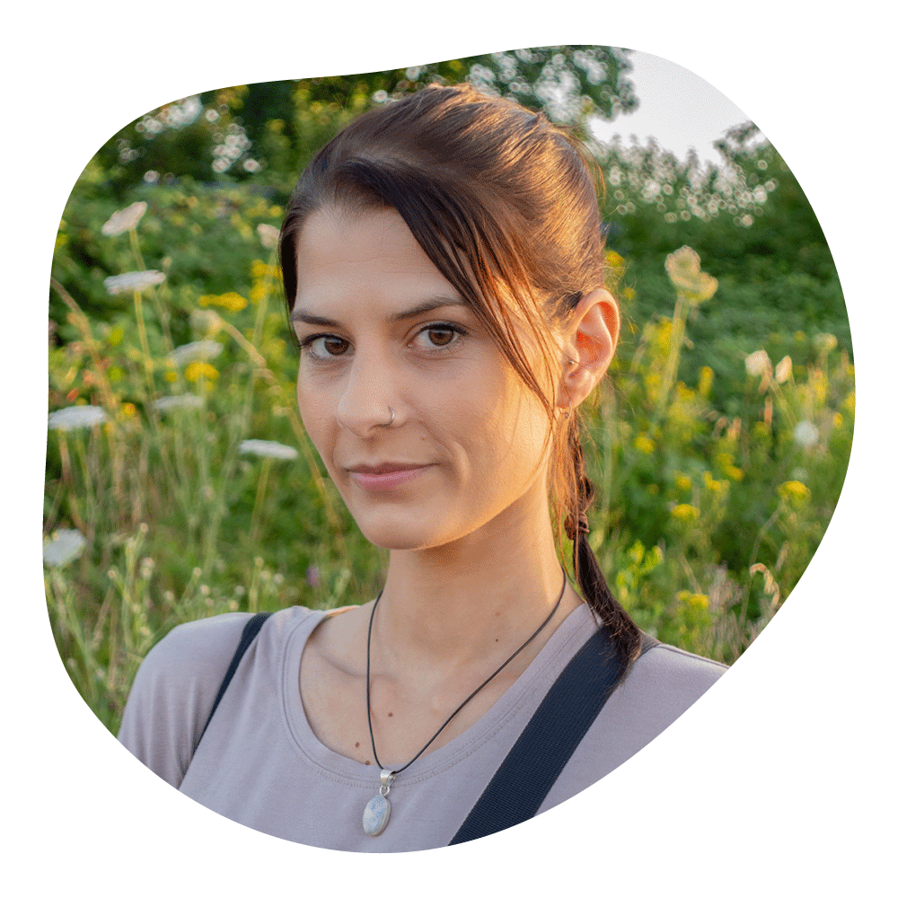 Junge Frau mit dunklen Haaren im Zopf, im Freien, umgeben von Blumen und Bäumen, Sonnenlicht auf dem Gesicht, trägt einen Beutel, eine Halskette mit einem Anhänger und ein T-Shirt.