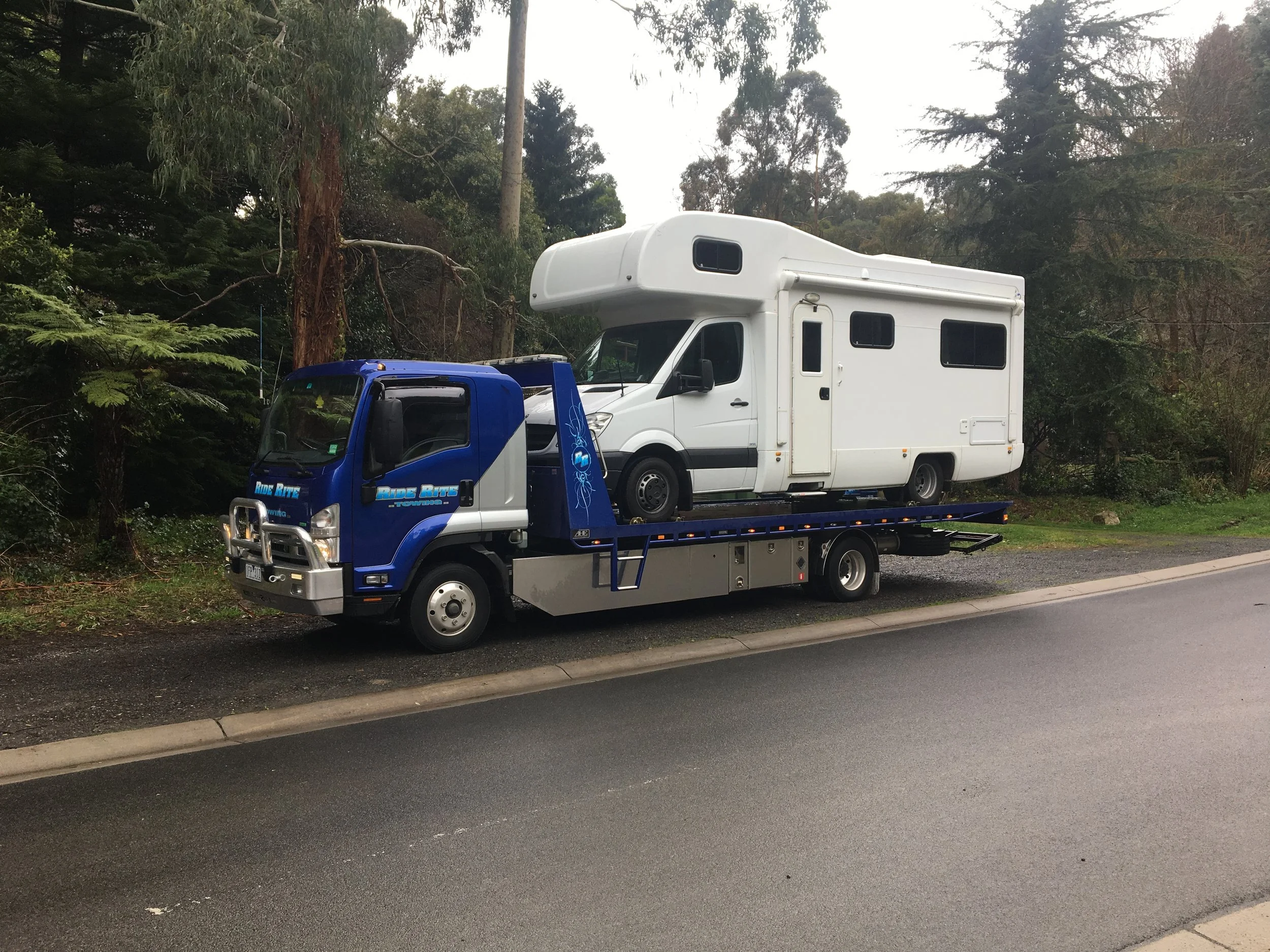A flatbed tow truck transporting a white camper van on a rural roadside.