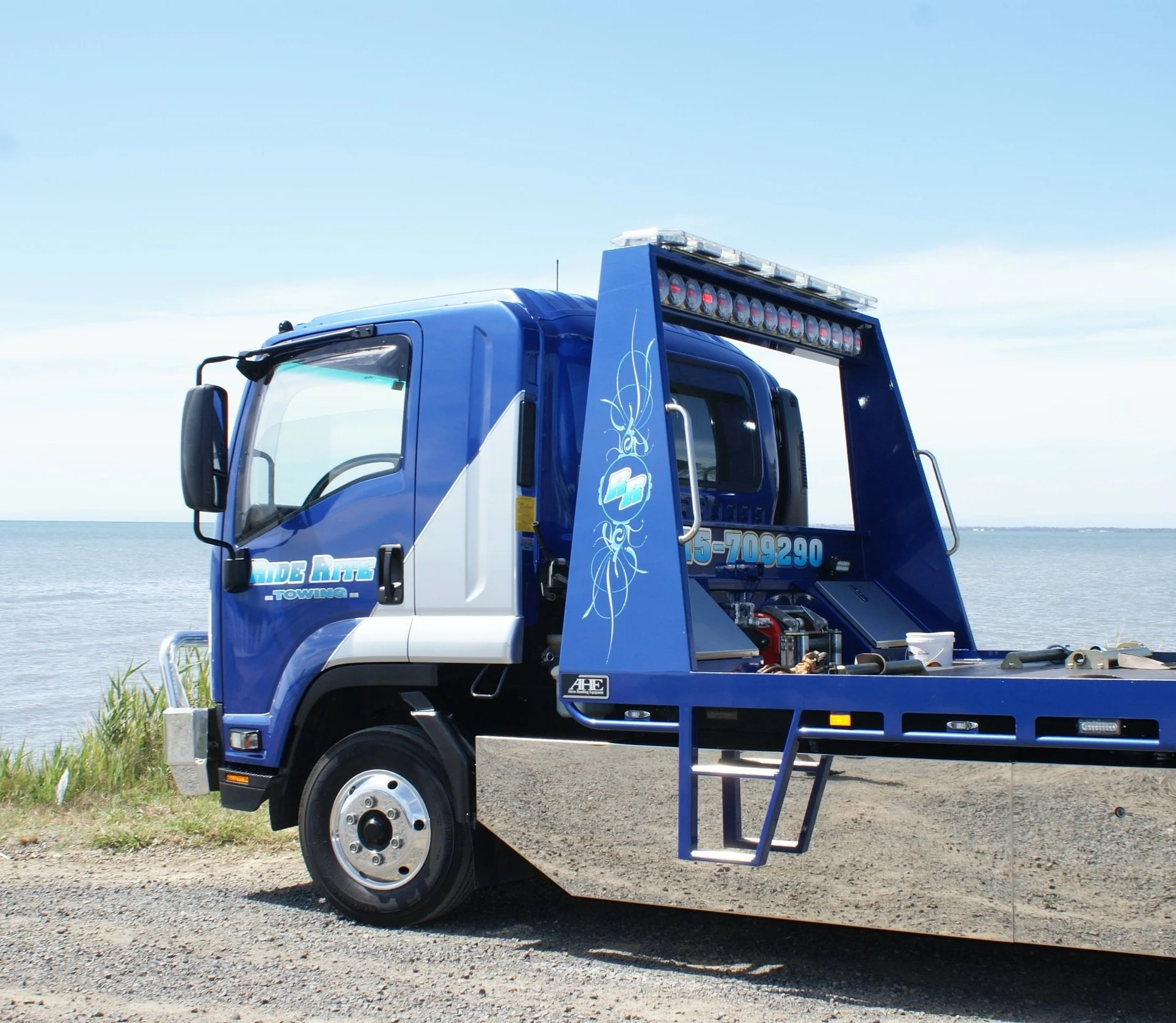Blue tow truck parked by the shoreline on a sunny day.