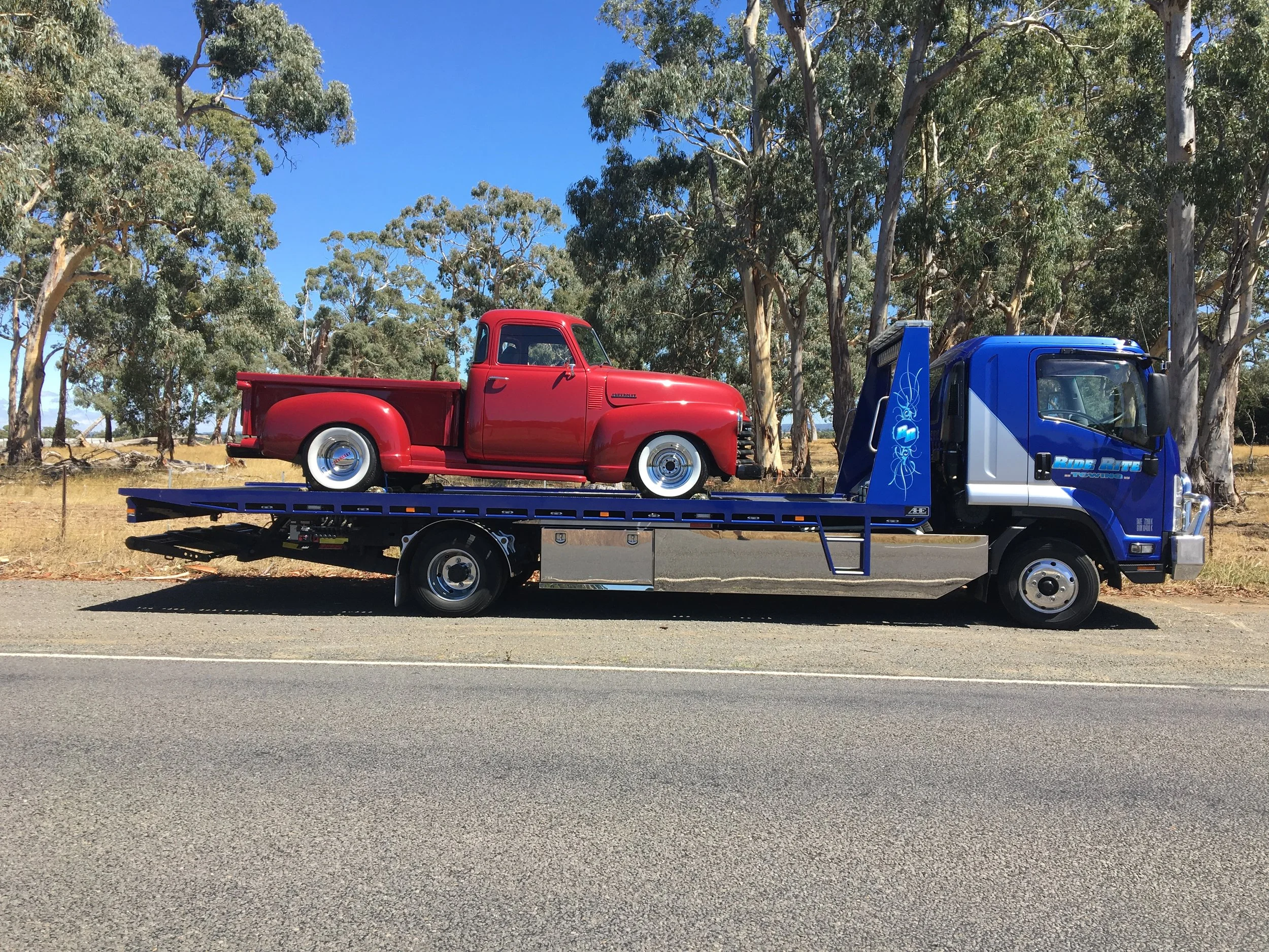 A red vintage pickup truck on a blue flatbed tow truck parked on a road, surrounded by eucalyptus trees.