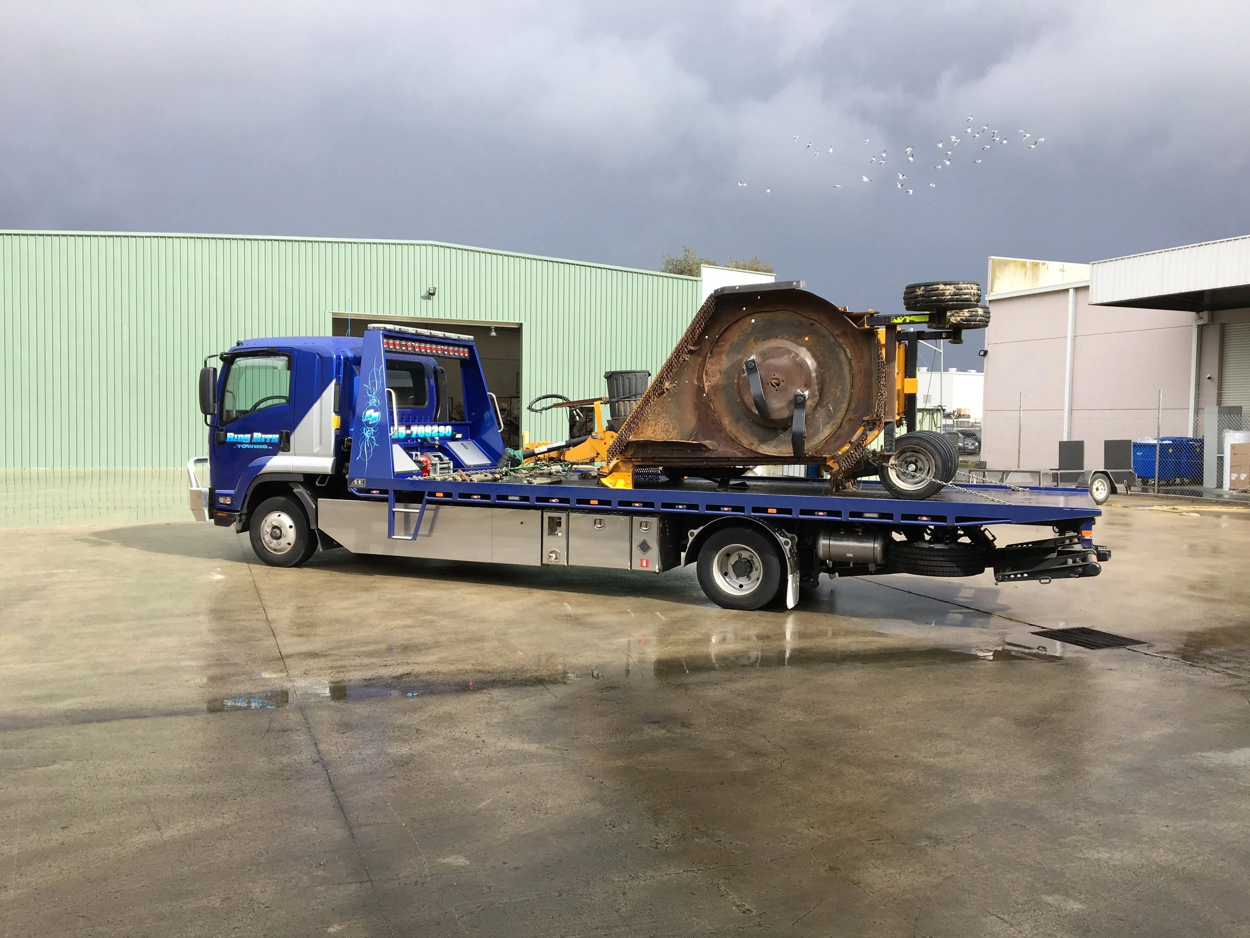 Blue tow truck carrying a large industrial machine part in an industrial yard.
