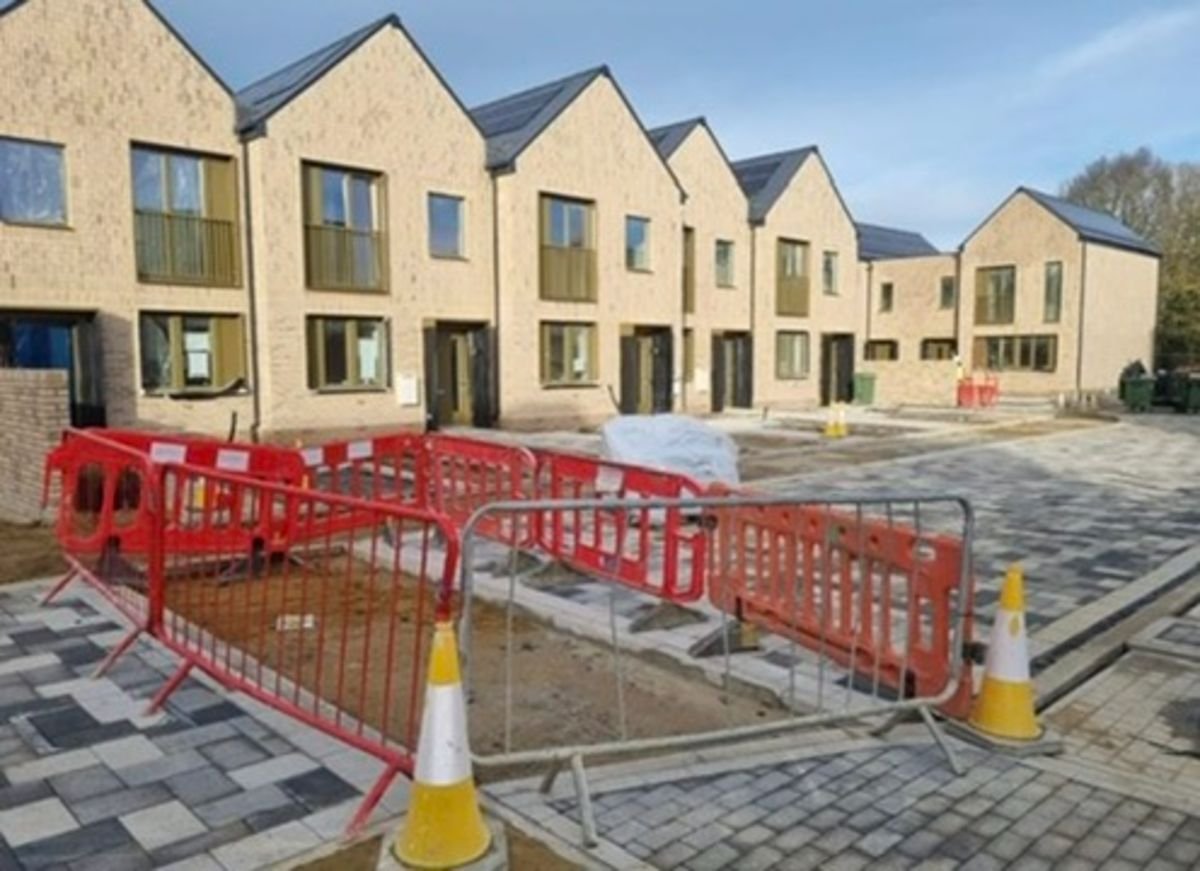 Newly constructed row of modern townhouse buildings with a partially paved courtyard area. Red plastic barriers and traffic cones indicate a construction zone in front of the houses.