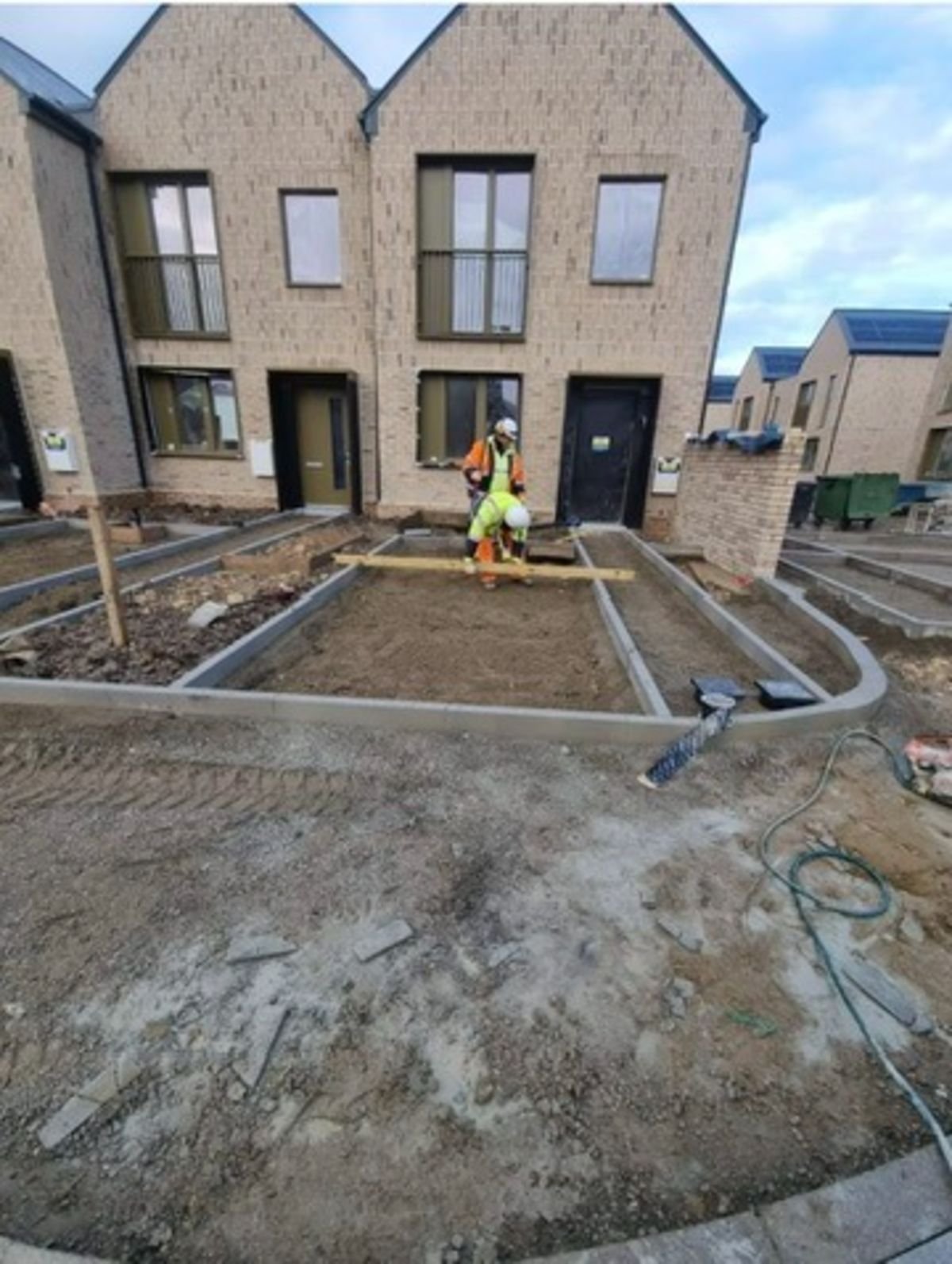 Two construction workers in high-visibility gear preparing a concrete foundation outside two-story brick houses under construction.