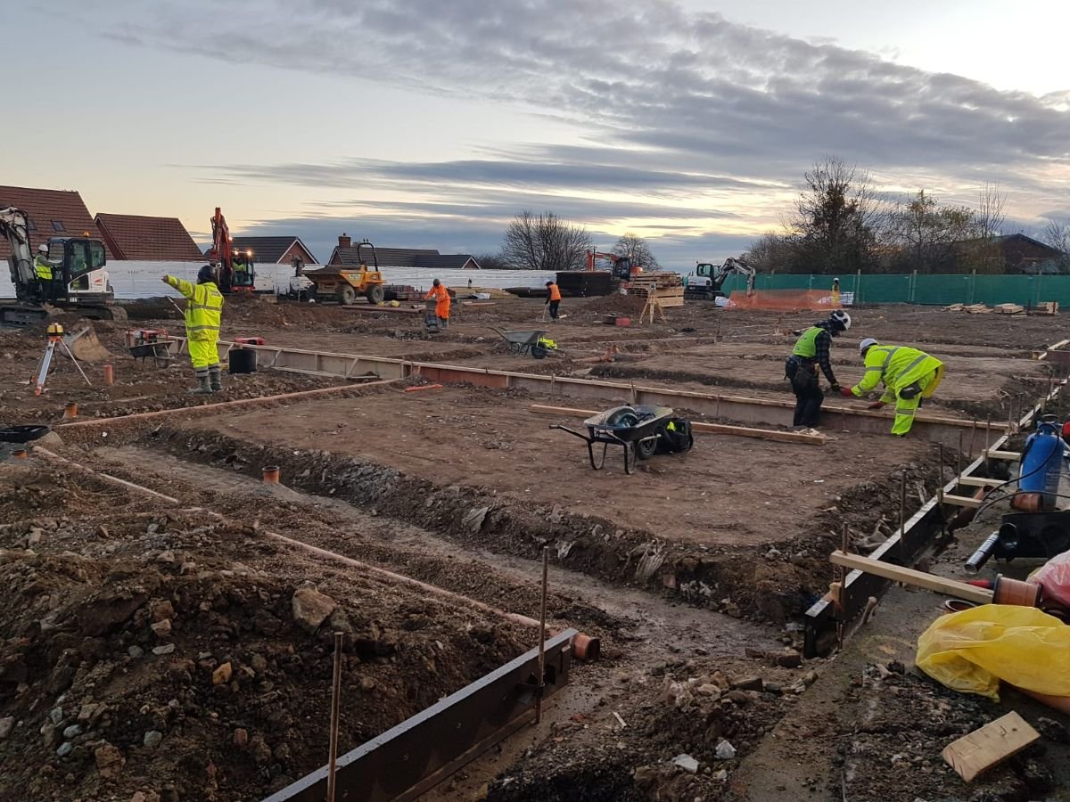 Construction site with workers in safety gear leveling ground and operating machinery. Equipment includes excavators, a dump truck, and a wheelbarrow. Residential houses in background, clear sky overhead.