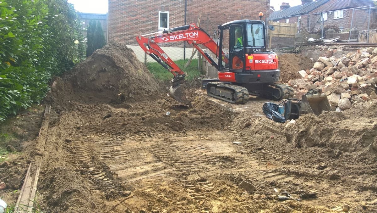 Construction site with an excavator, pile of dirt, and rubble. It is set near residential buildings, with a hedge on one side and a brick wall in the background.