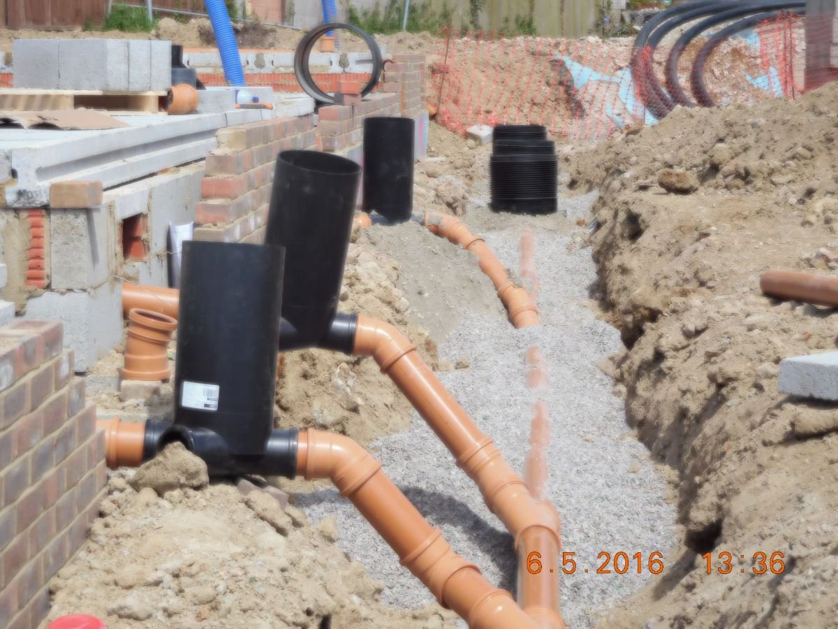 Construction site showing drainage pipes, brickwork, and gravel in a trench.