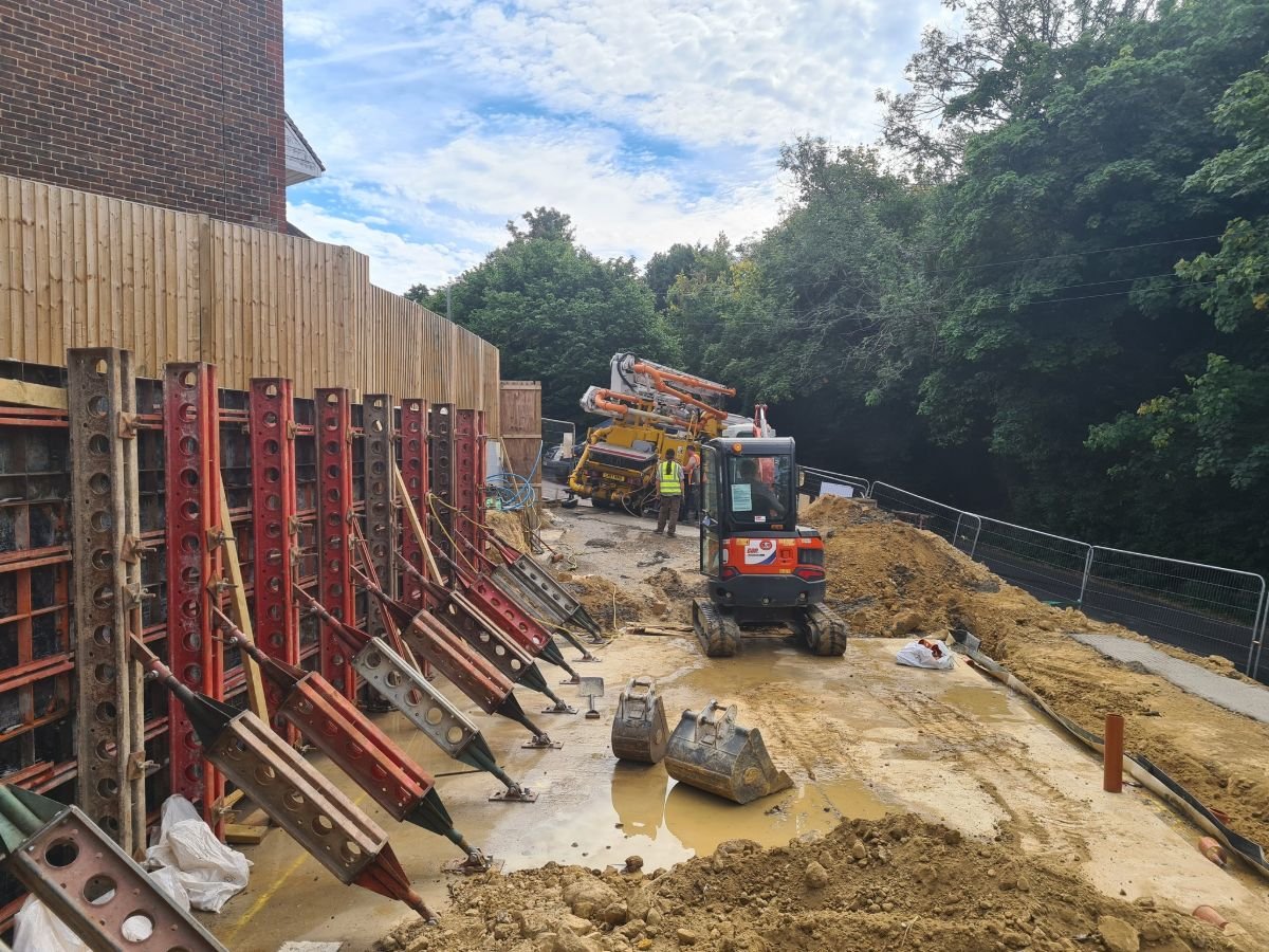 Construction site with heavy machinery, including a small excavator and concrete mixer truck. Wooden fencing and metal supports are visible, along with excavated earth and construction materials.