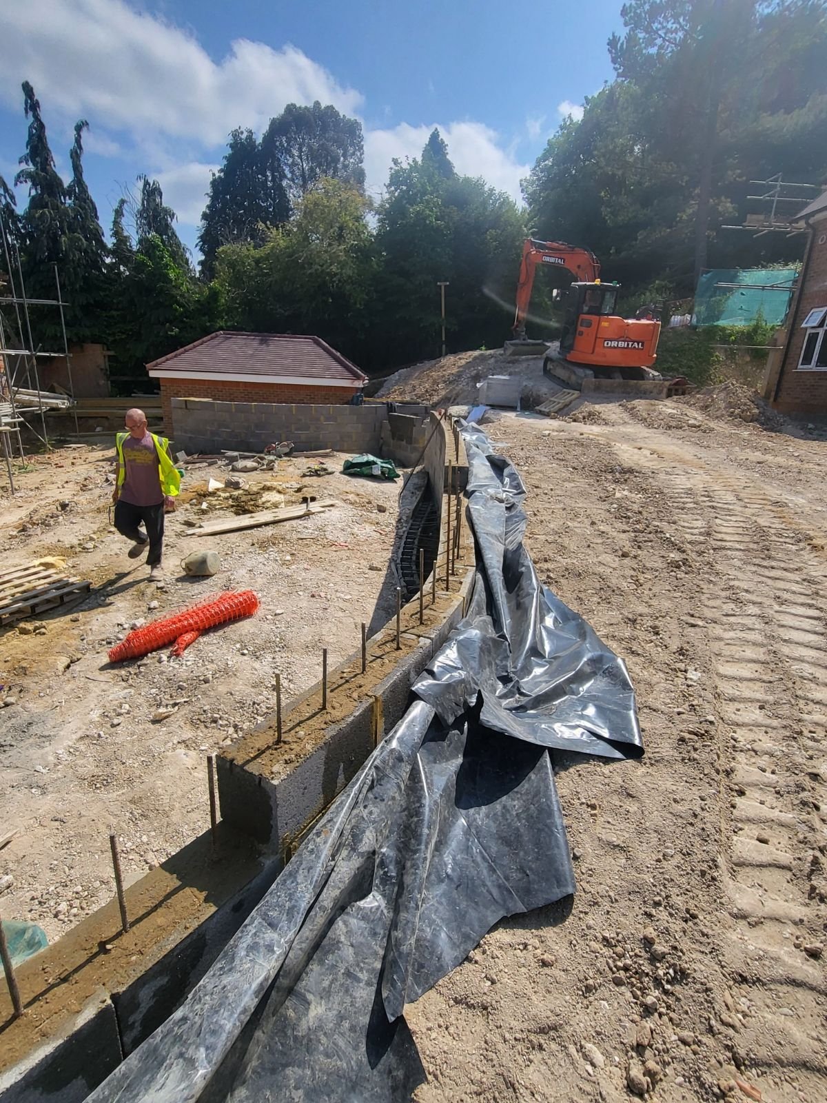 Construction site with a partially built wall, an excavator, a person in a high-visibility vest, and various building materials. Trees and a small building are in the background.