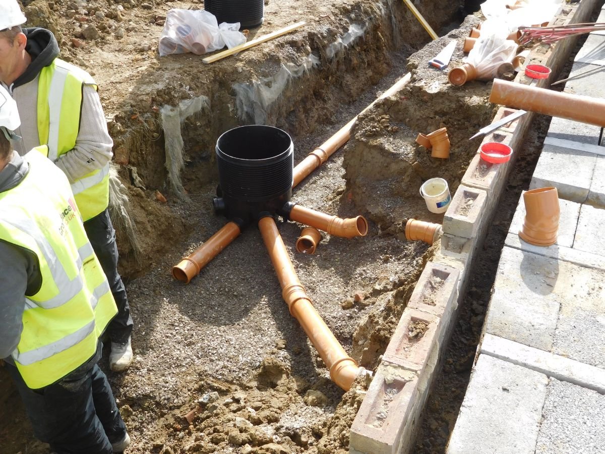 Construction site with workers installing underground pipes near a trench. Visible materials include orange drainage pipes, connectors, and construction tools. Workers are wearing safety helmets and high-visibility vests.