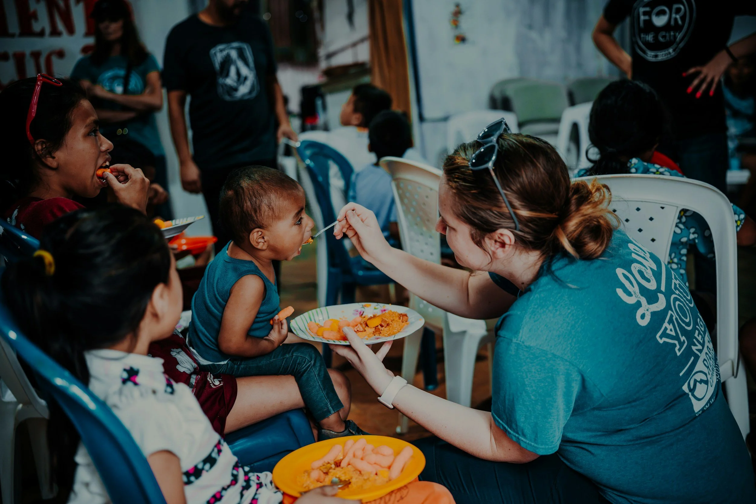 A woman feeds a young child with a spoon at a gathering, with children sitting nearby and others in the background.