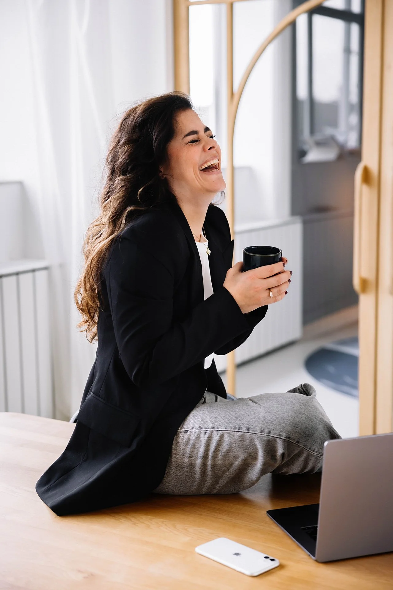 Een lachende vrouw zit op een tafel met haar benen op de tafel, houdt een zwarte mok vast en kijkt naar rechts. Ze draagt een zwarte blazer en een witte top, met een grijze broek. Op de tafel liggen een laptop en een witte smartphone.