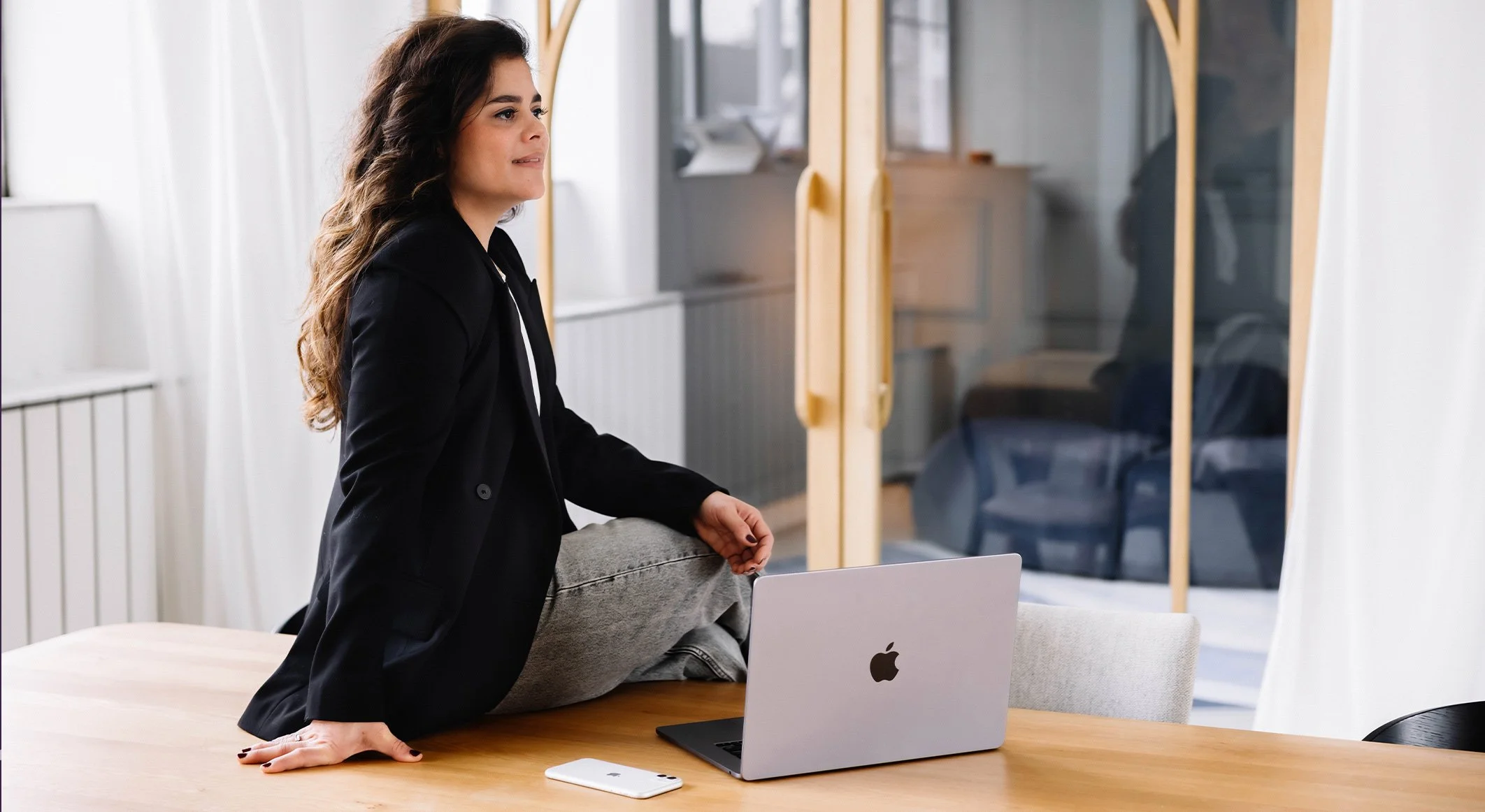 Vrouw zit op de tafel met één been, naast een open MacBook en een witte smartphone in een lichte kamer met glazen deuren.