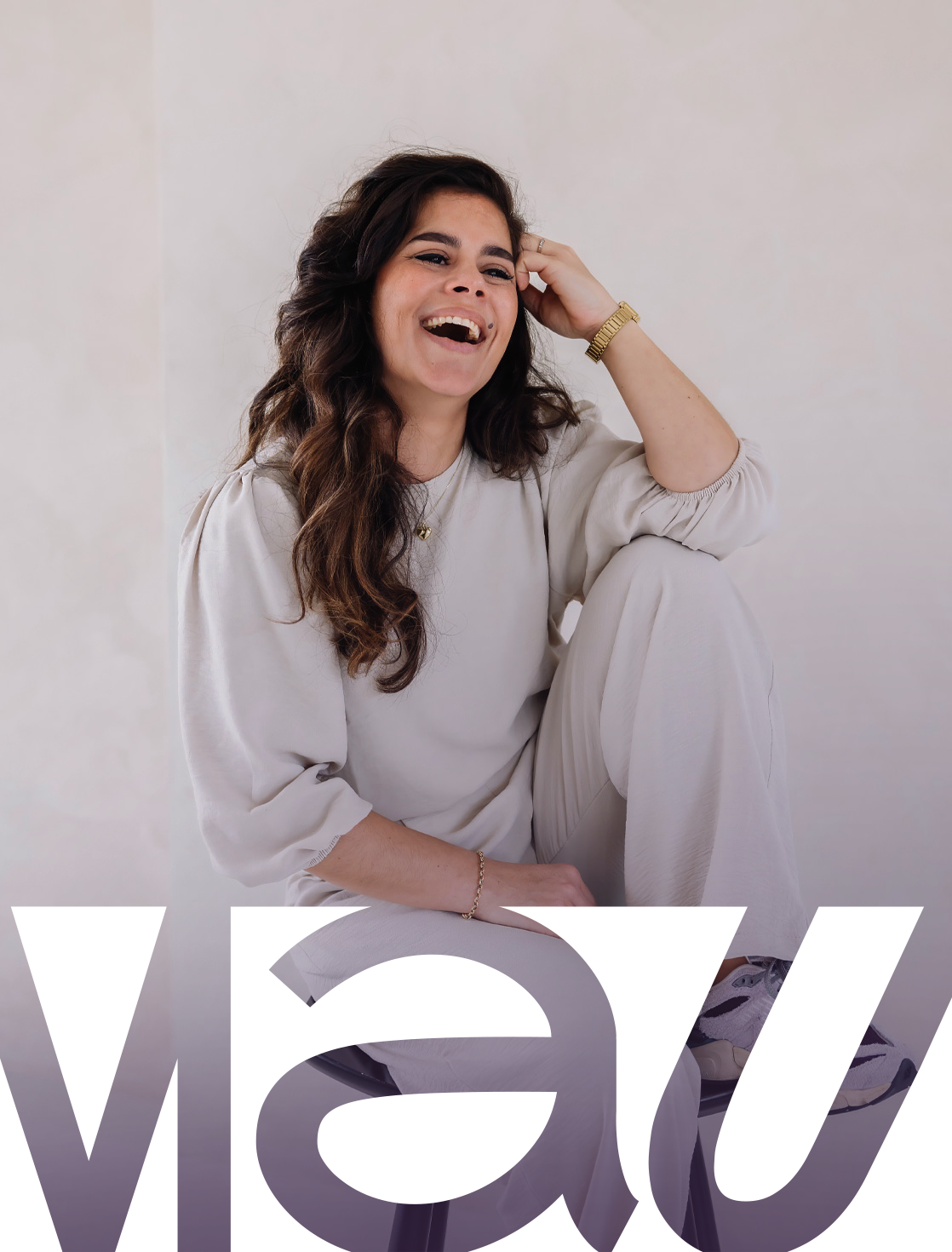 Een lachende vrouw met lang, donkerbruin haar, gekleed in een crèmekleurig shirt en broek, zittend op een stoel, lachend en met haar hand in haar haar, witte achtergrond.