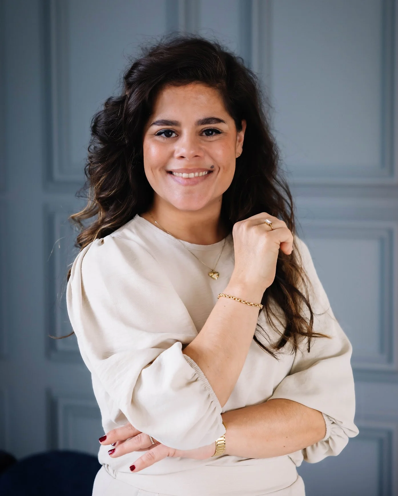 Vrouw met bruin, golvend haar die glimlacht en poseren in een lichte blouse, met een grijze deur op de achtergrond.