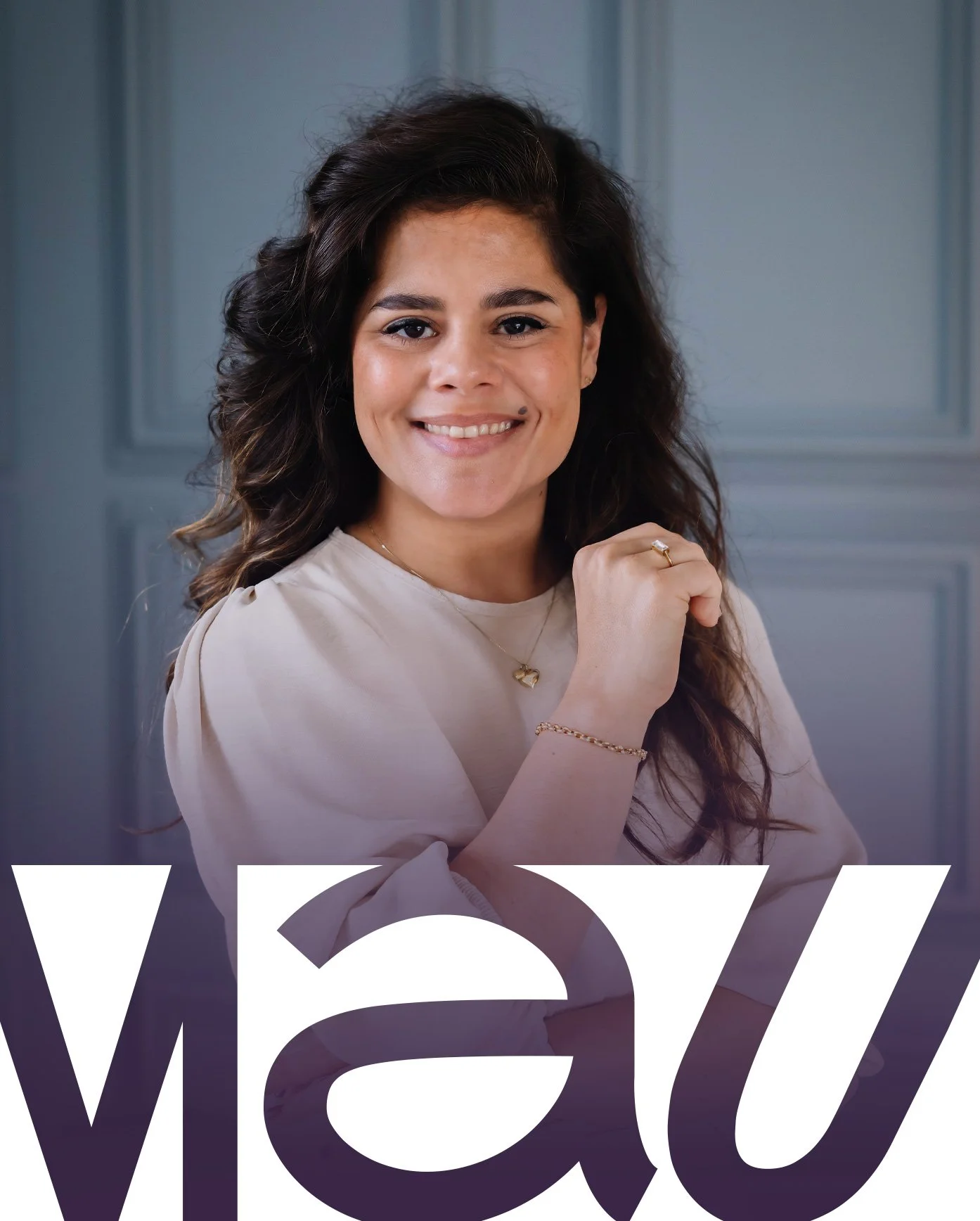 Vrouw met lange haar en glimlach, gekleed in een lichtkleurige blouse, draagt sieraden en zit voor een blauwe achtergrond met grote tekst die gedeeltelijk zichtbaar is.