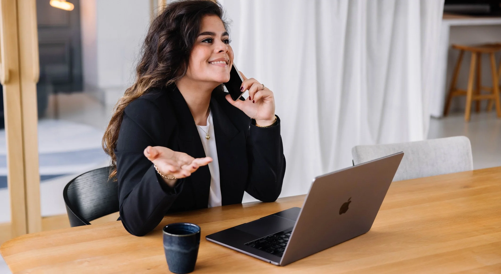 Vrouw zit aan een houten tafel, heeft een laptop en een kopje, en praat op de telefoon terwijl ze een verwarde of ongeduldige uitdrukking toont.