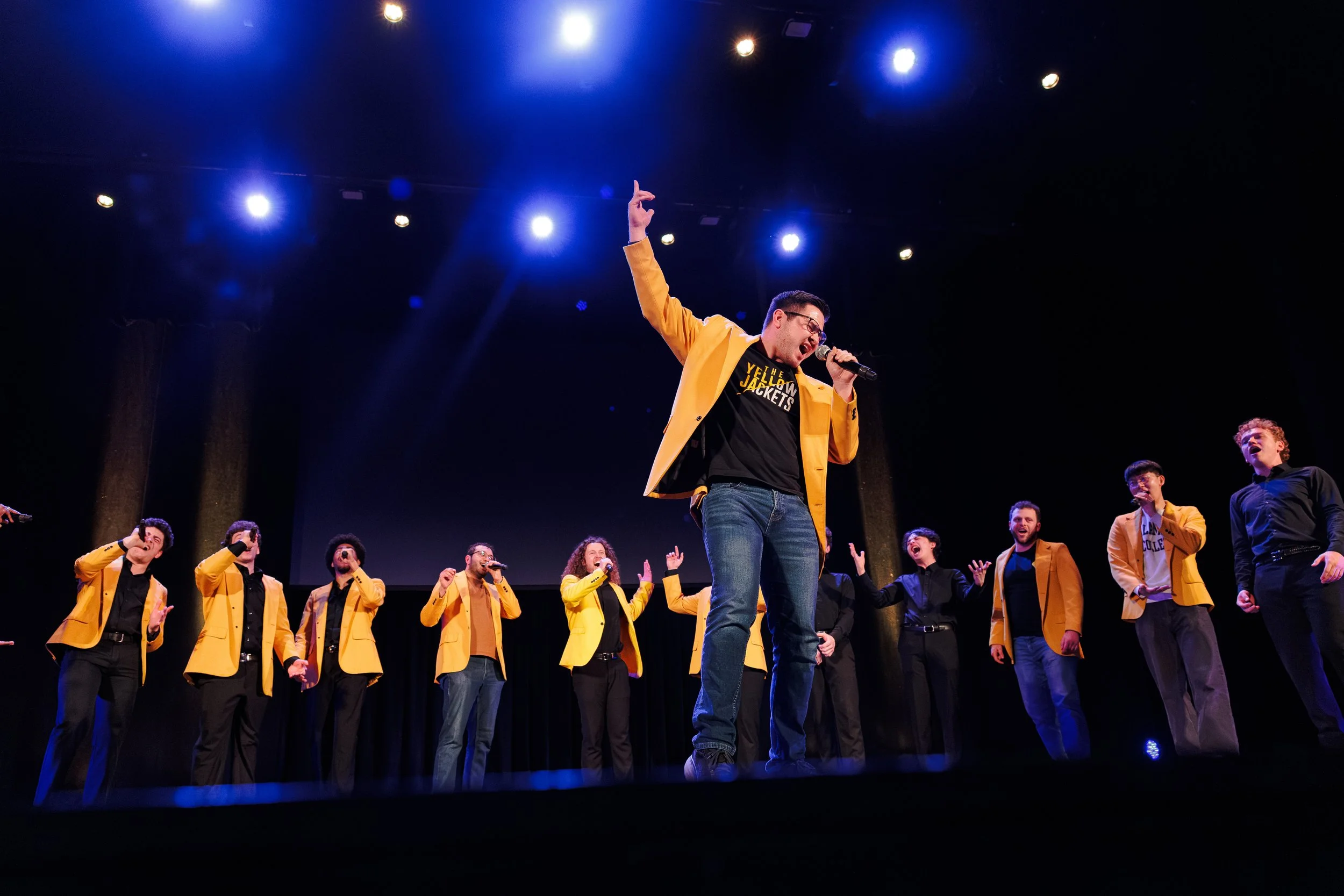 A group of performers on stage, with one man in a yellow jacket singing into a microphone and others clapping and cheering, under bright stage lights.