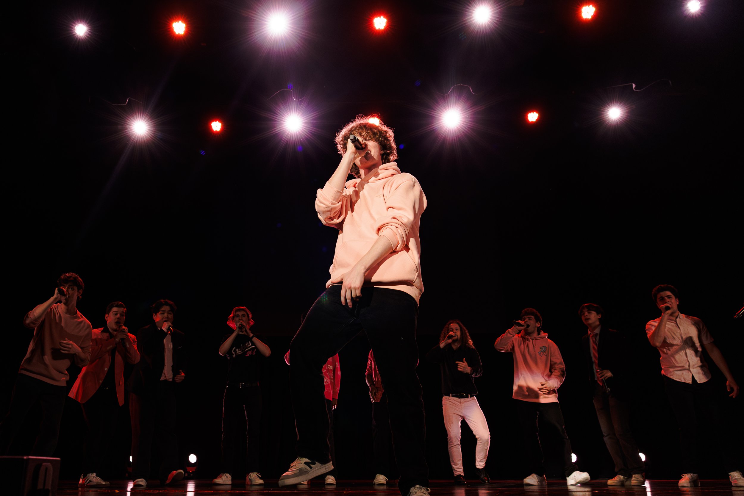 A young performer with curly hair and a peach hoodie singing into a microphone on stage illuminated by bright spotlights, surrounded by a group of young people.