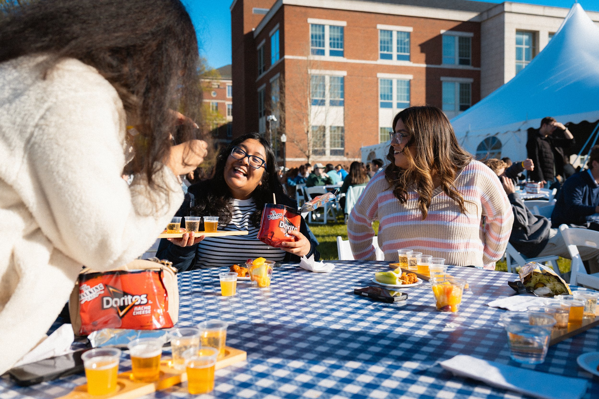 People gathering outdoors, enjoying food and drinks at a daytime event with a blue checkered tablecloth, in front of a brick building and a white tent.