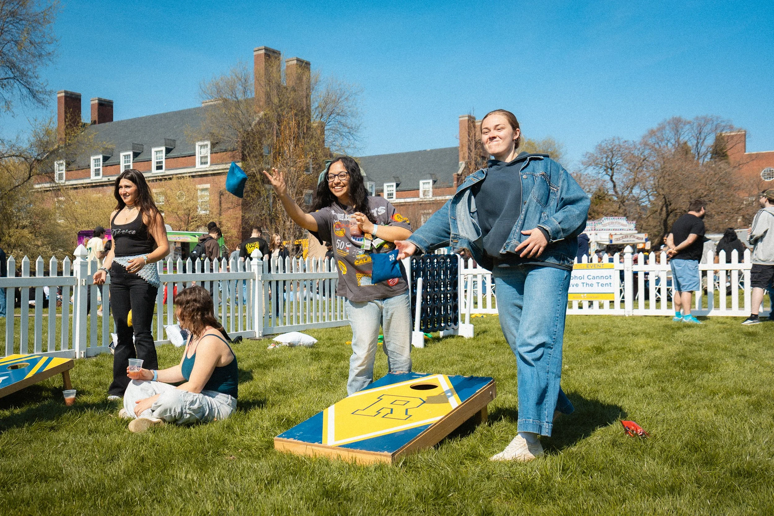 Three young women playing cornhole outdoors on a sunny day. Two are standing and tossing bean bags, one is sitting on the grass watching. There are other people and food trucks in the background, with a large brick building and trees.