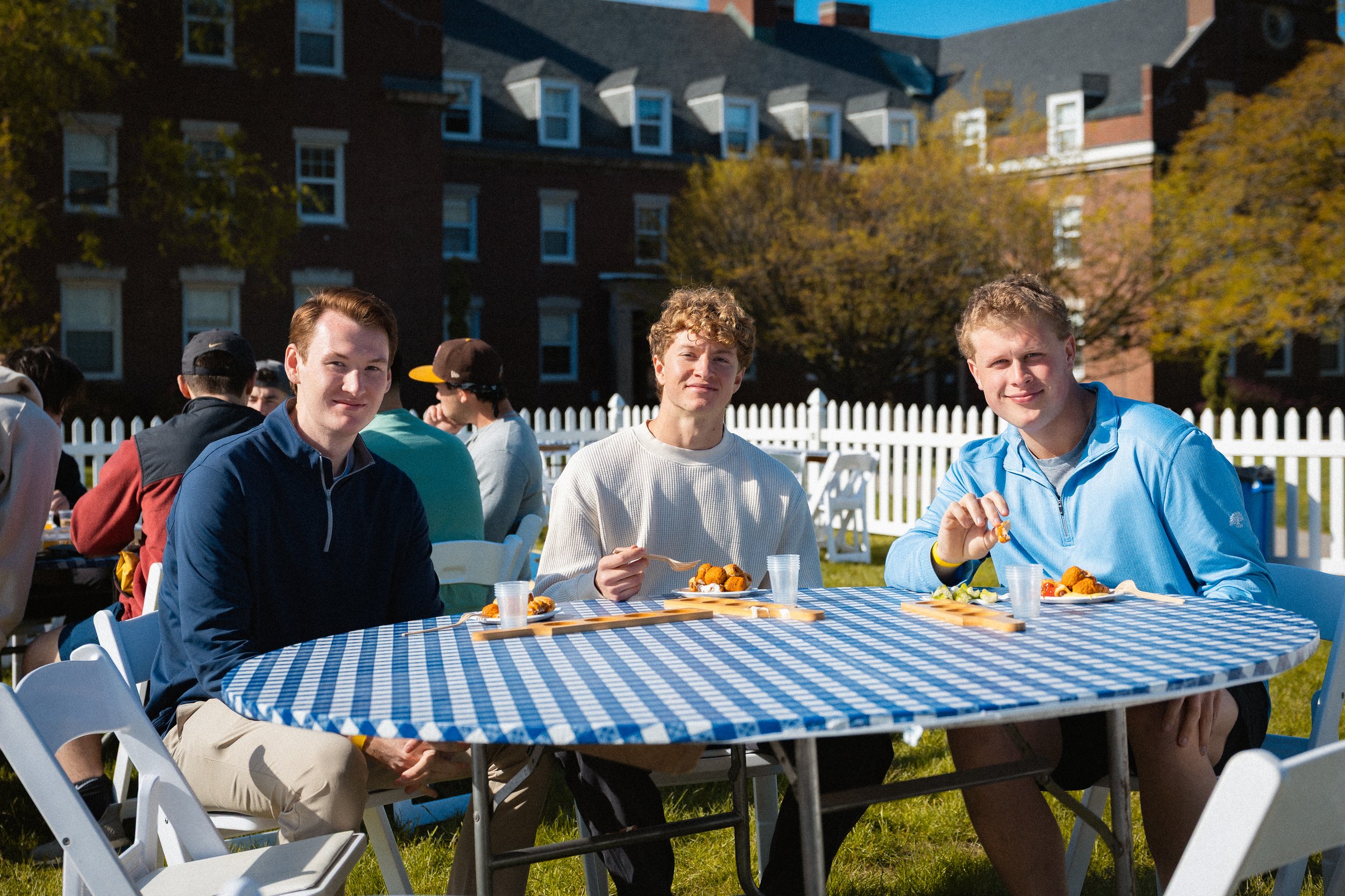Three young men sitting at a checkered blue and white outdoor table, enjoying food with a group of people in the background during a sunny day, with a brick building and trees behind them.