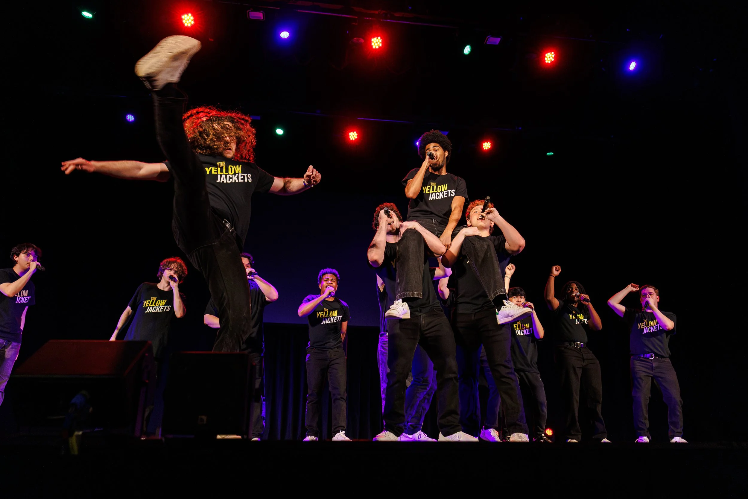 Group of young performers on stage wearing matching black t-shirts with yellow text, some holding microphones, engaged in singing or performing, with colorful stage lights overhead.