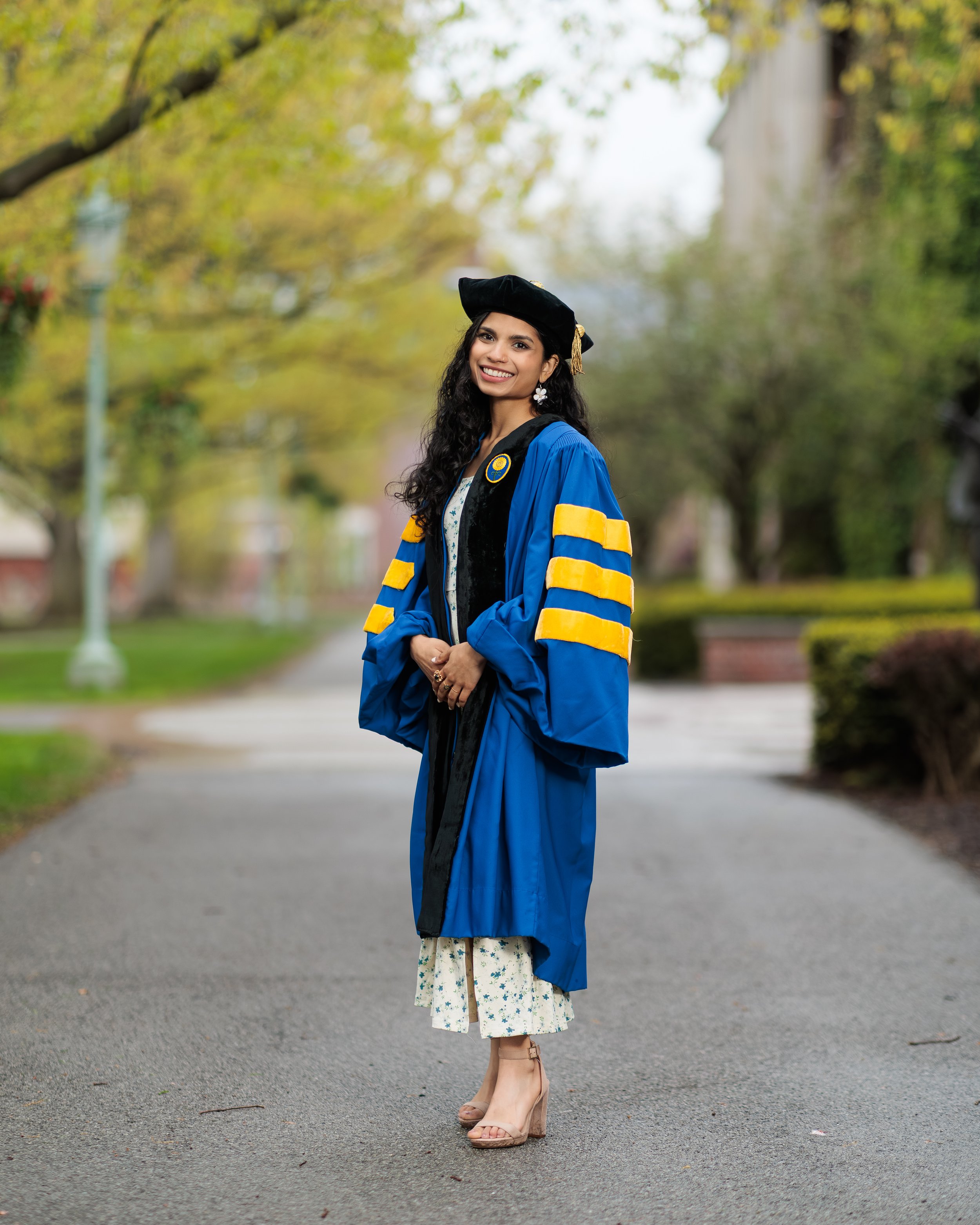 A young woman in graduation attire standing outdoors on a paved walkway, surrounded by trees with green and yellow leaves, smiling at the camera.