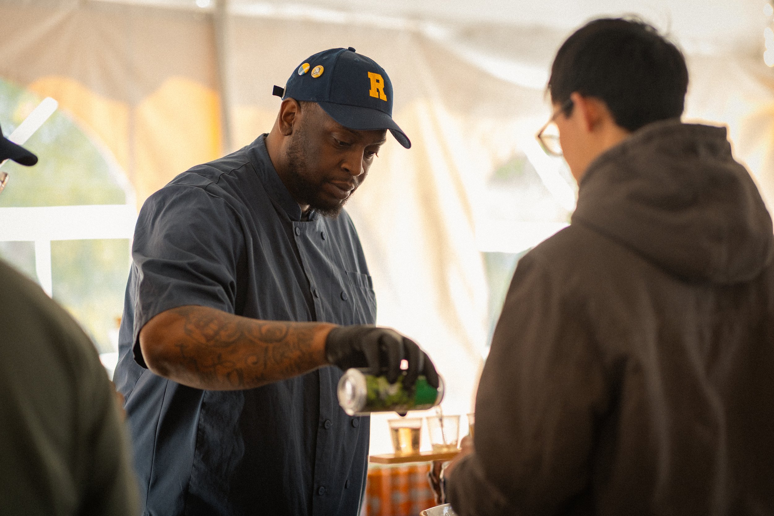 A man wearing a navy baseball cap with a yellow 'R' and buttons, gray chef's coat, and gloves working in a kitchen, with another person in the foreground wearing glasses and a dark hoodie.