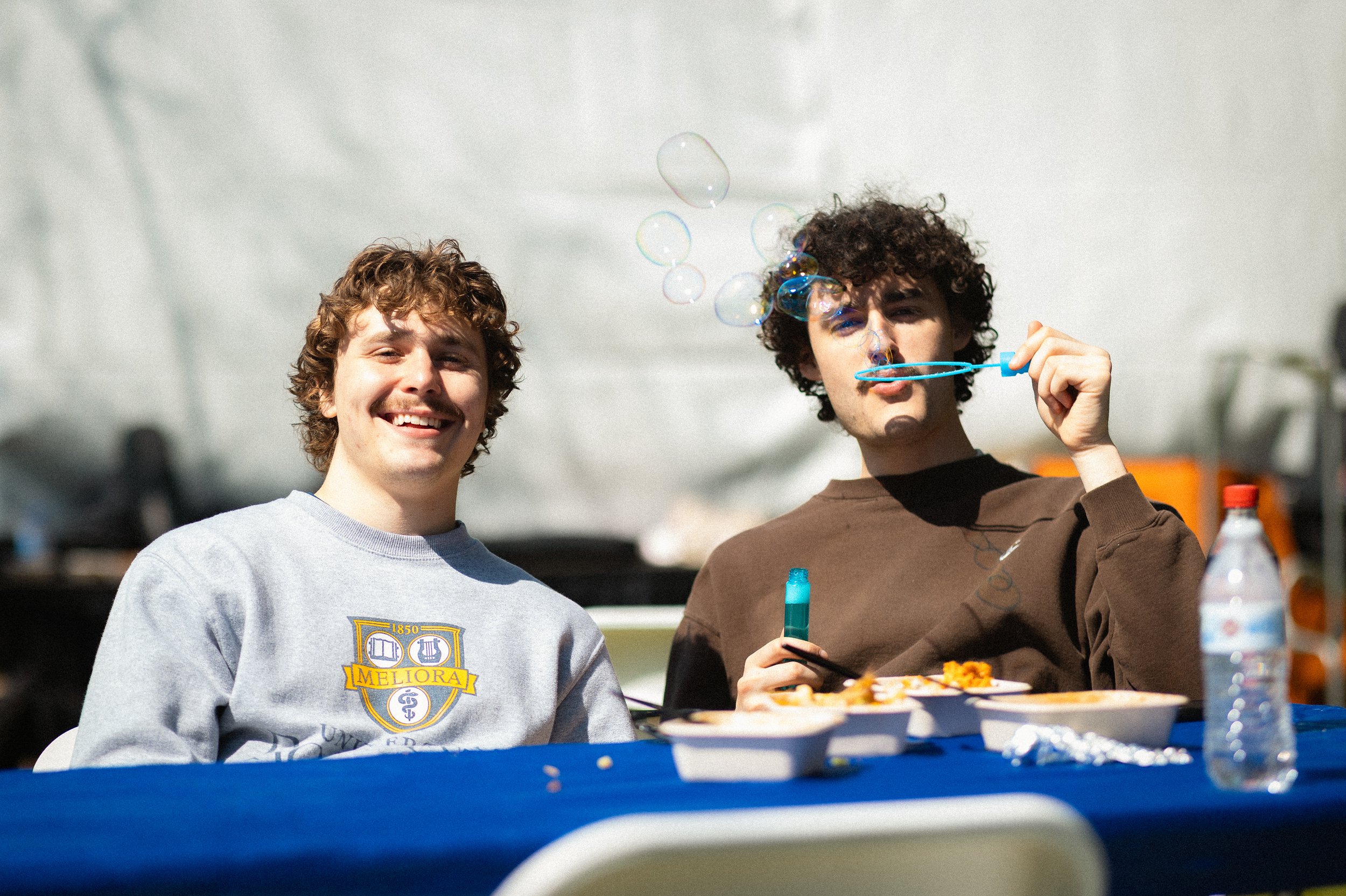 Two young men sitting at a table outdoors. One is smiling at the camera while the other is blowing soap bubbles. Food and drinks are on the table.