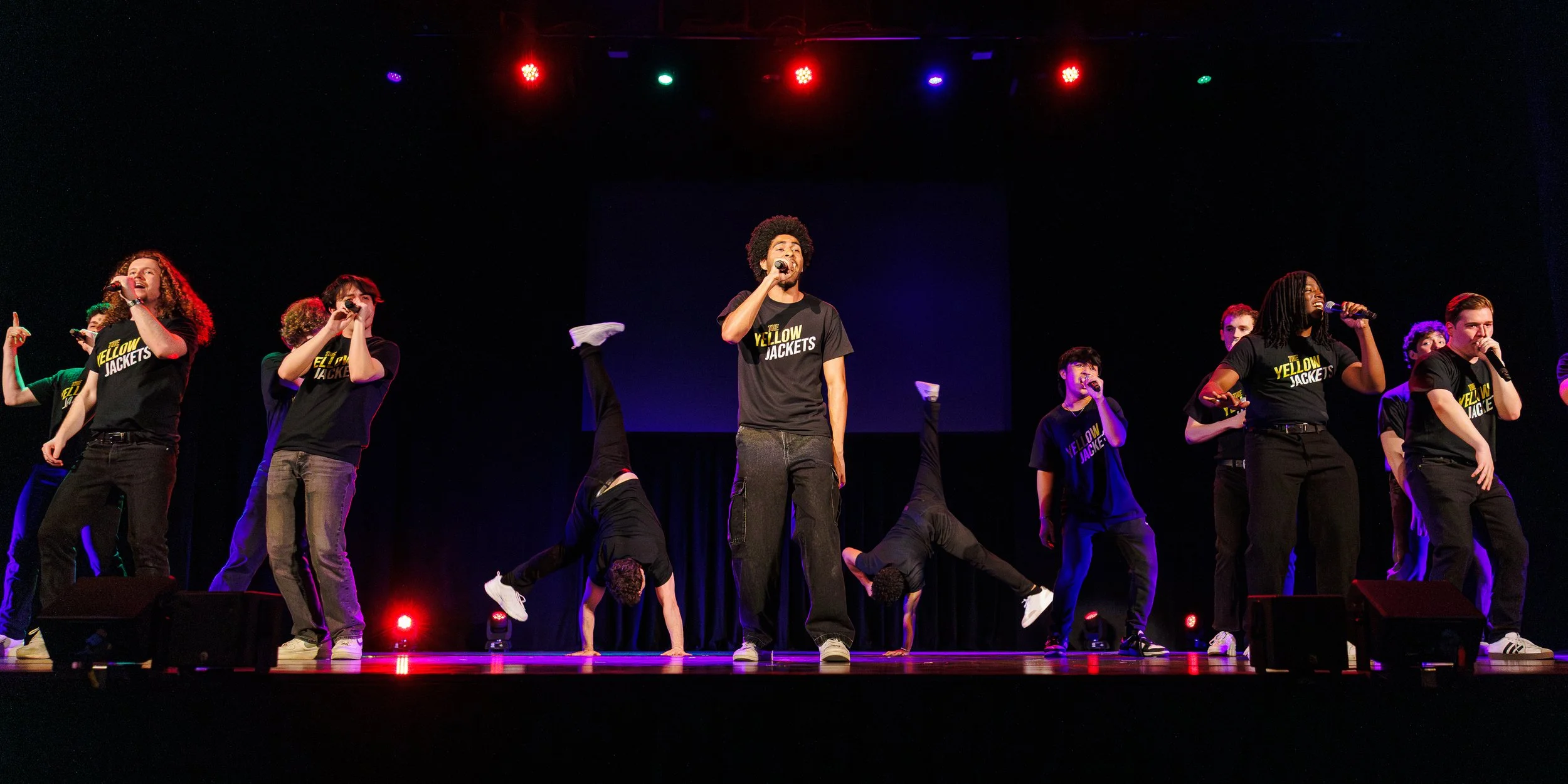Group of young performers on stage, some singing into microphones, others doing handstands, all wearing matching black T-shirts with yellow text reading 'The Yellow Jackets', under colorful stage lights.