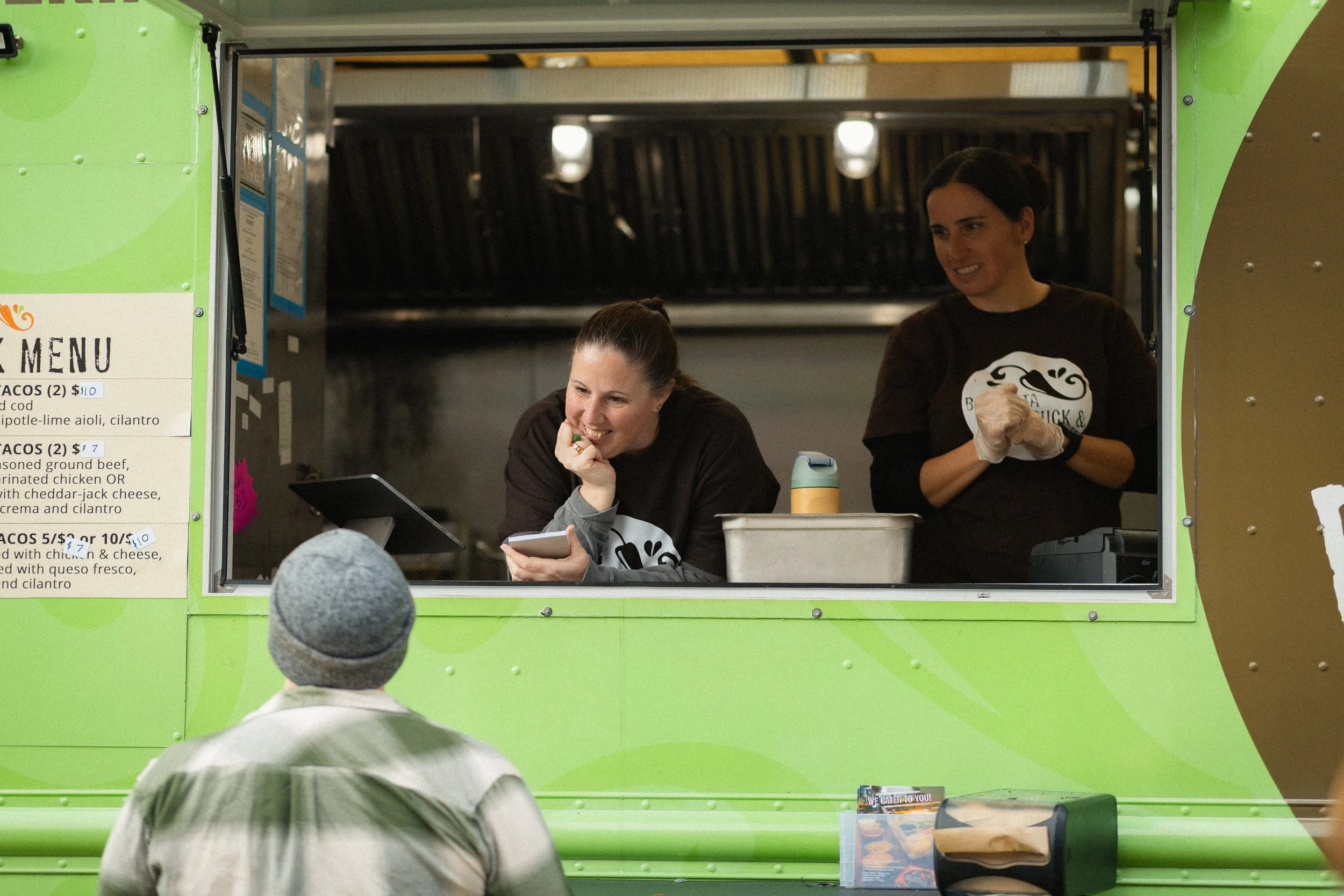 Two women working at a green food truck window, interacting with a customer. One woman is smiling and looking at a tablet, the other is smiling and wearing gloves. A customer wearing a gray beanie is standing outside.