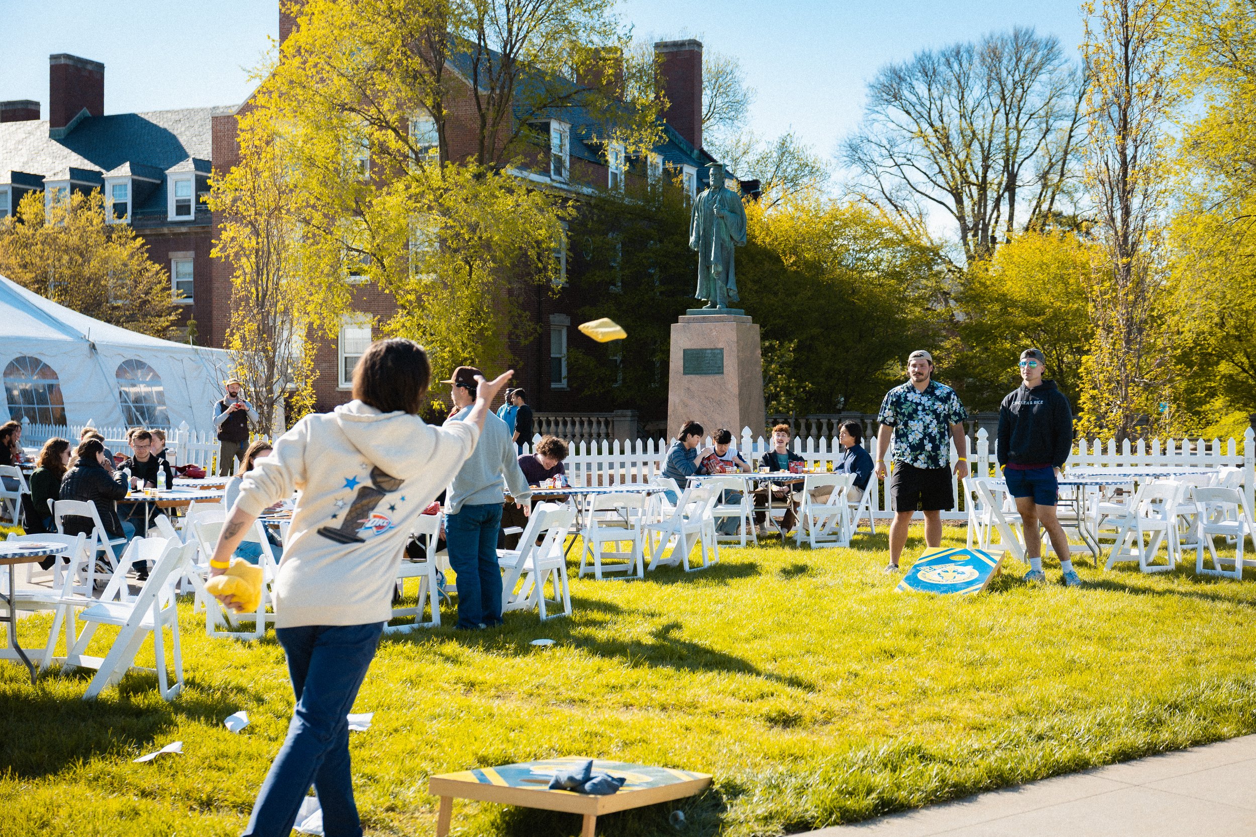 People gathered outdoors at a daytime event with white chairs, some throwing paper, in front of a statue and trees with yellow leaves.