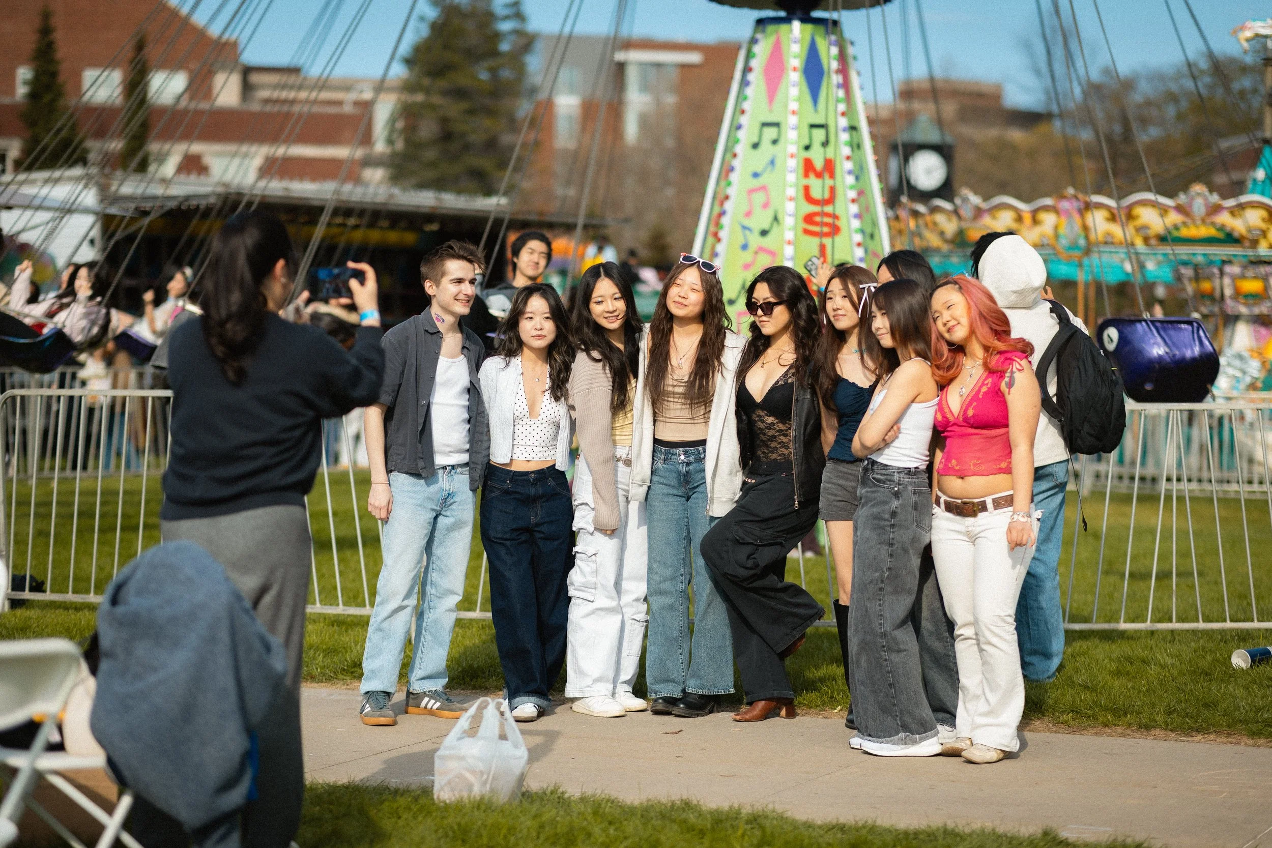 A group of diverse young people posing for a photo at a carnival or fairground with a colorful amusement ride in the background.