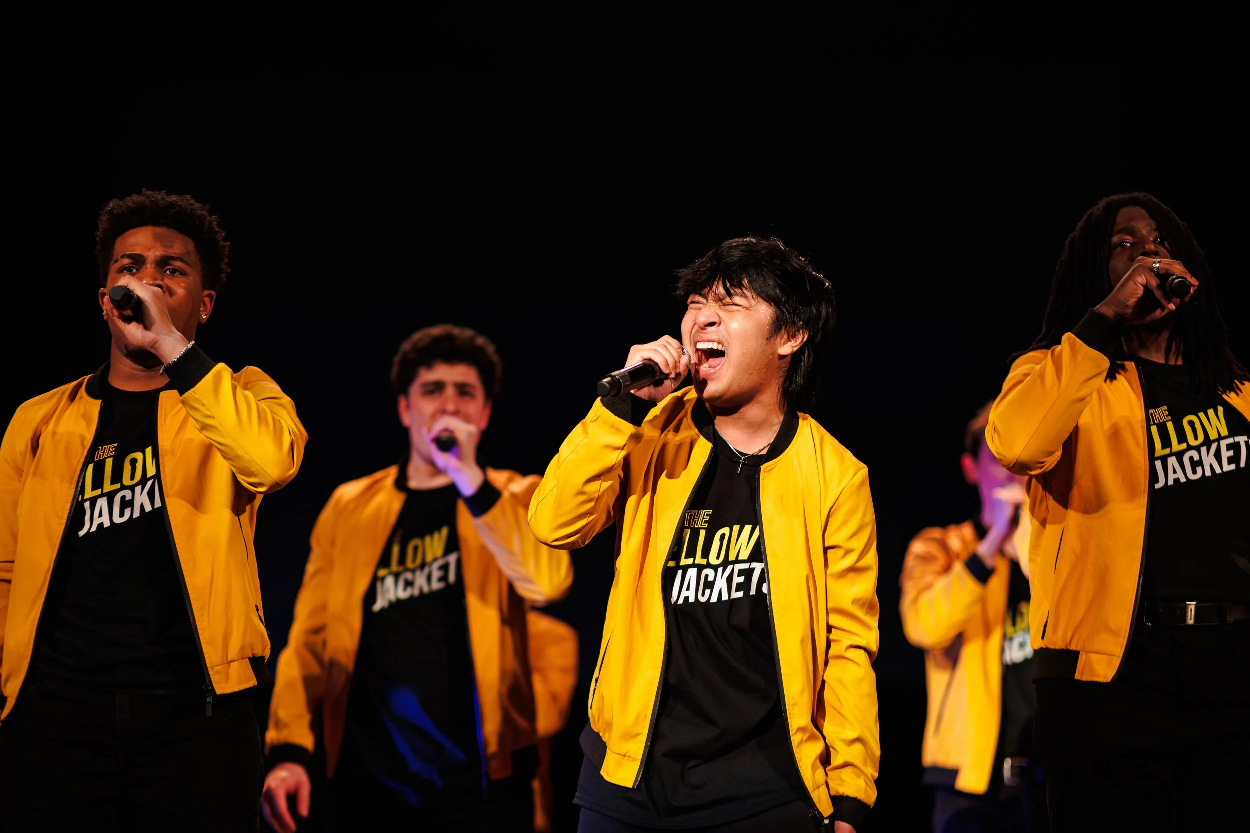 Group of people singing into microphones, wearing yellow jackets and black shirts with yellow text, on stage with black background.