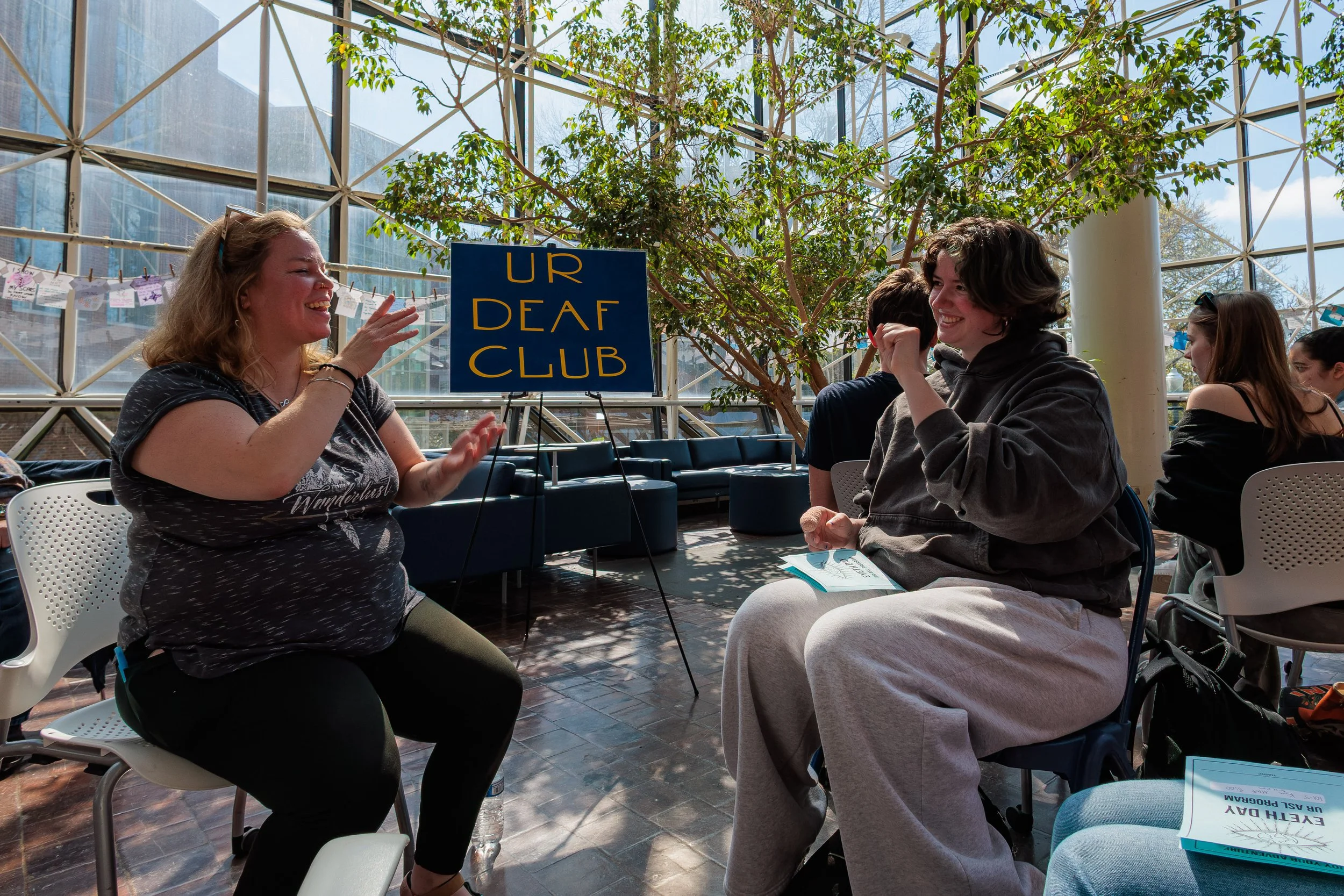 A group of people sitting and chatting at a table inside a glass-enclosed conservatory with a sign that reads 'UR DEAF CLUB'.