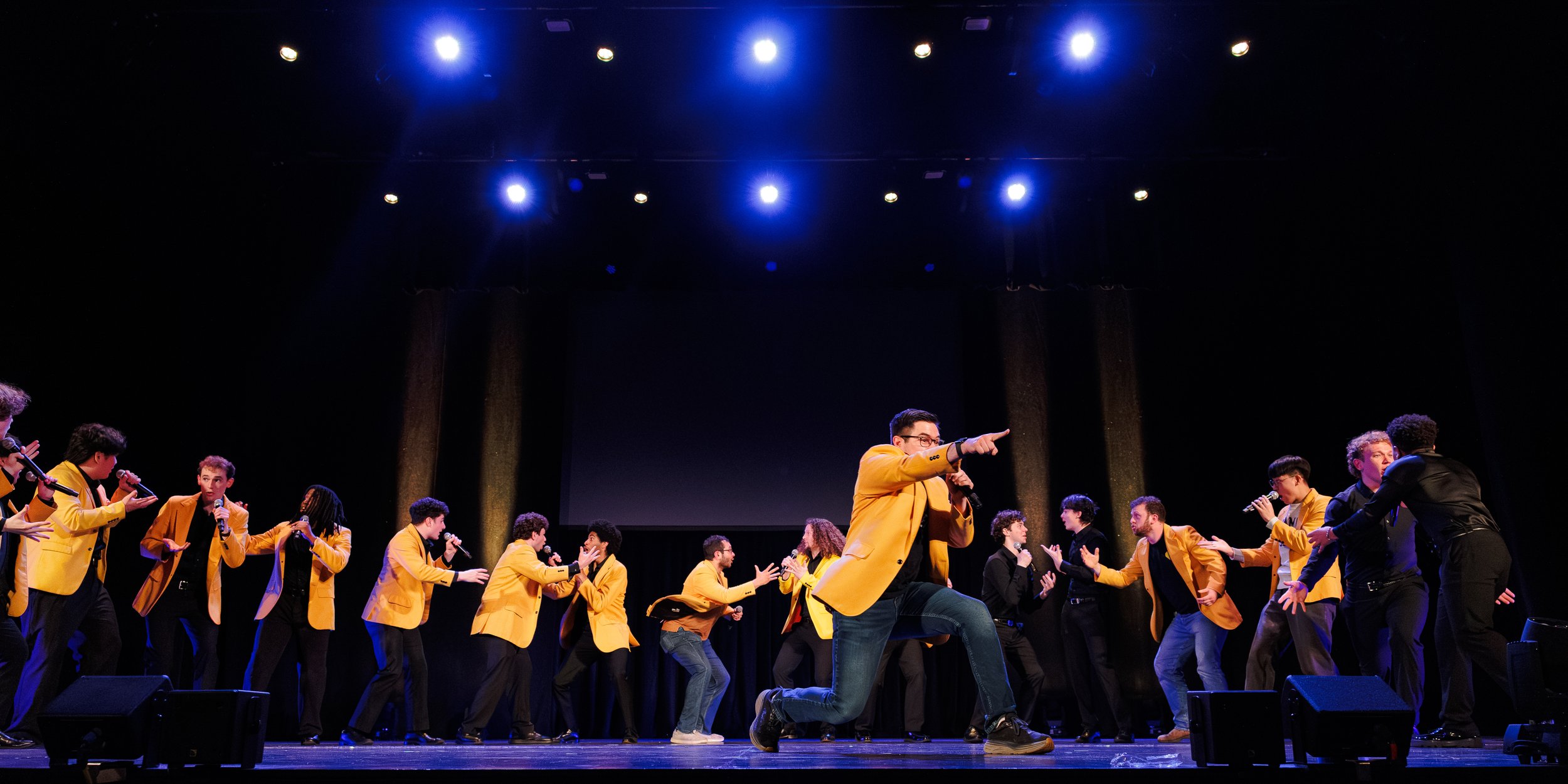 A group of people performing on stage, many wearing yellow jackets, with one kneeling in front, all holding microphones and engaging with each other.