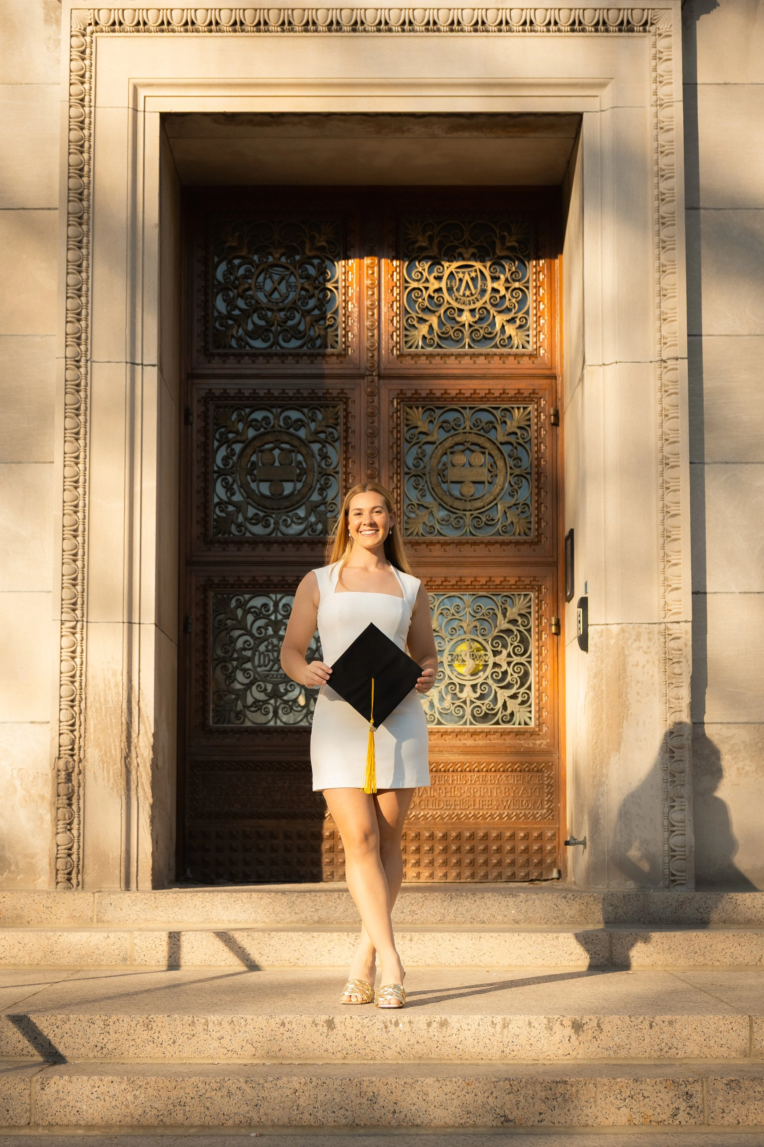 A young woman in a white dress holding a graduation cap, standing on the steps in front of an ornate large wooden door with intricate metalwork, smiling at the camera.