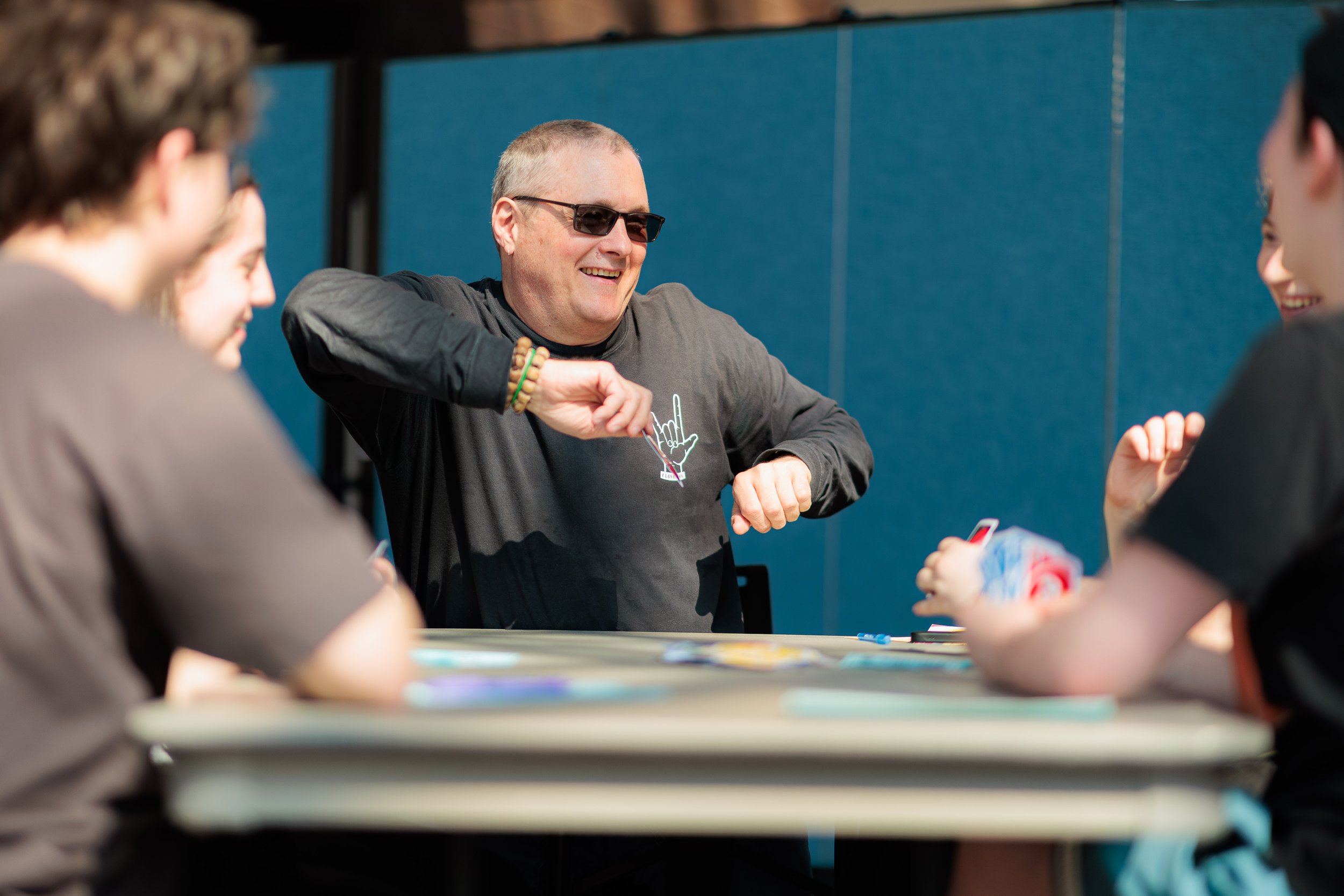 A man wearing sunglasses and a dark long-sleeve shirt smiles and interacts with a group of children sitting around a table, possibly playing a game or engaging in an activity in a room with a blue wall.
