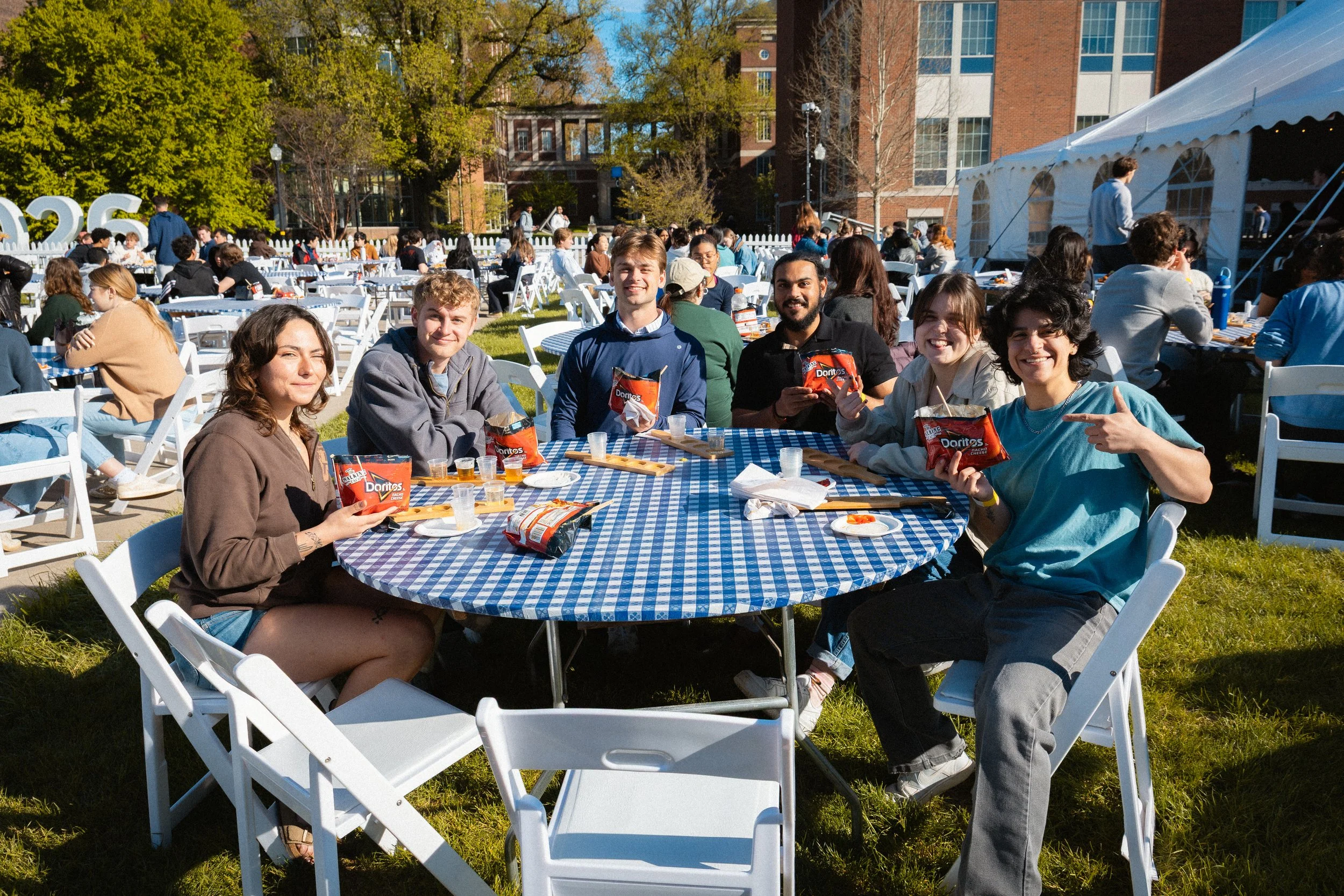 A group of young adults sitting at a round table outdoors, holding bags of Doritos, at a sunny event with many other people dining at similar tables, and a white tent in the background on a grass lawn with trees and a brick building.