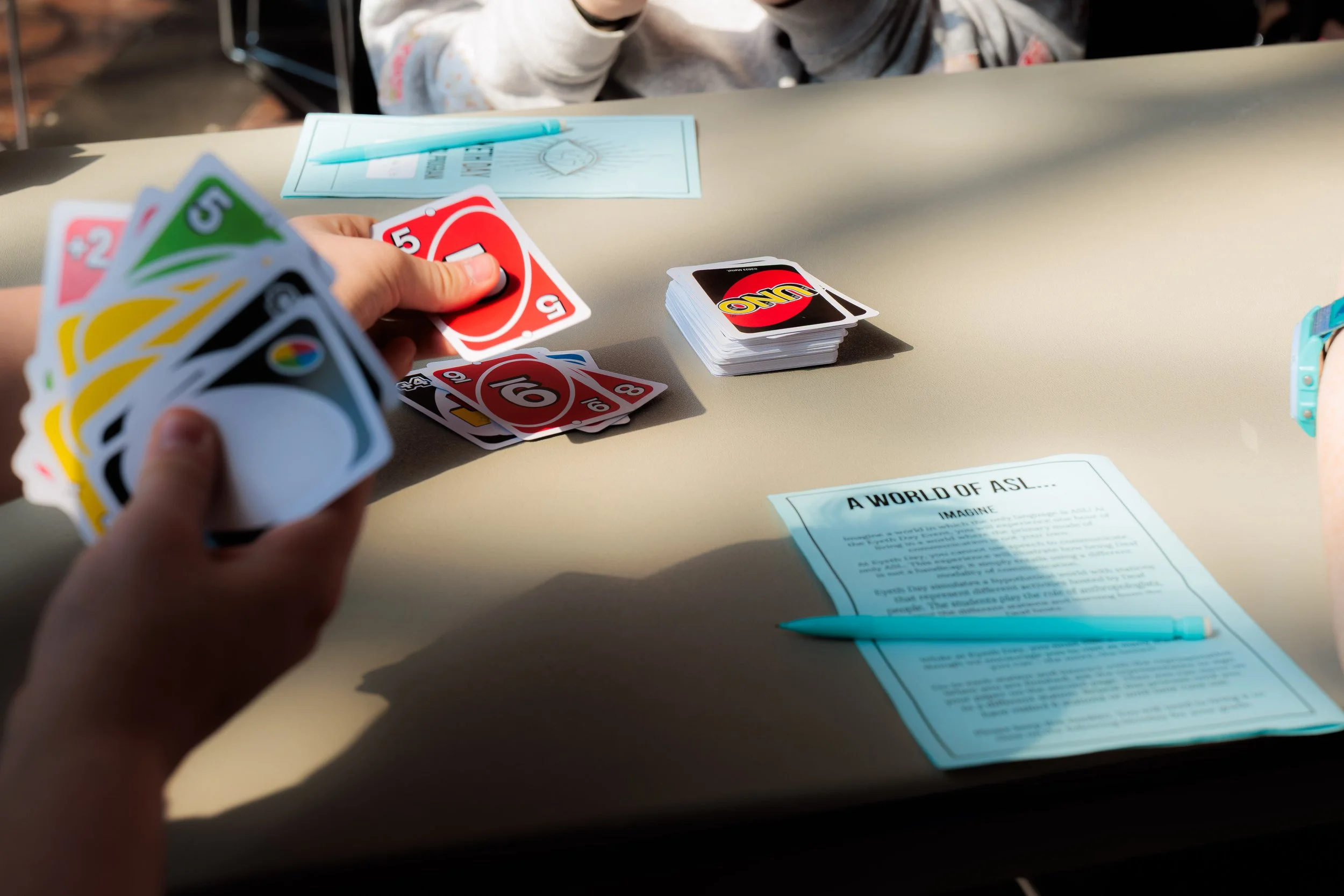 People playing Uno card game on a table with papers, pens, and wristbands.
