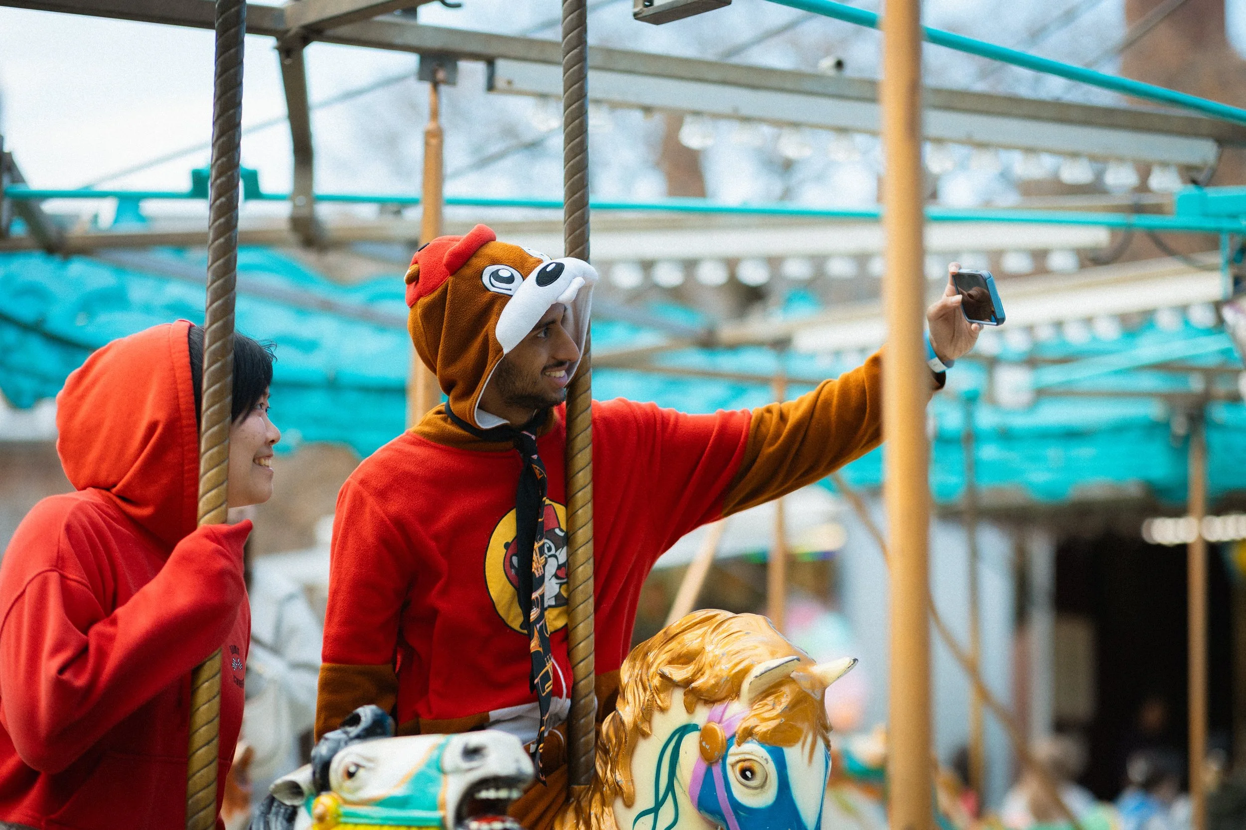 Two people, a woman and a man, are on a carousel with animal figures. The man is smiling and wearing a hoodie with a lion mascot, and the woman is also smiling, wearing a red hoodie. The man is taking a selfie with his phone.