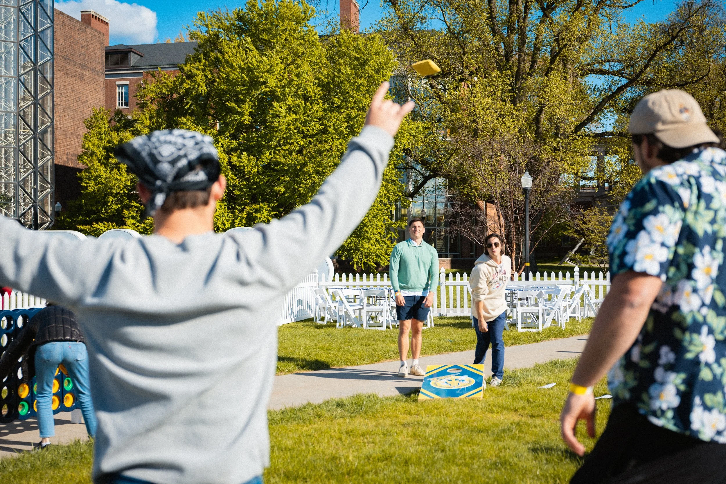Group of young adults playing a giant Connect Four game outdoors on a sunny day, with trees, a brick building, and white chairs in the background.