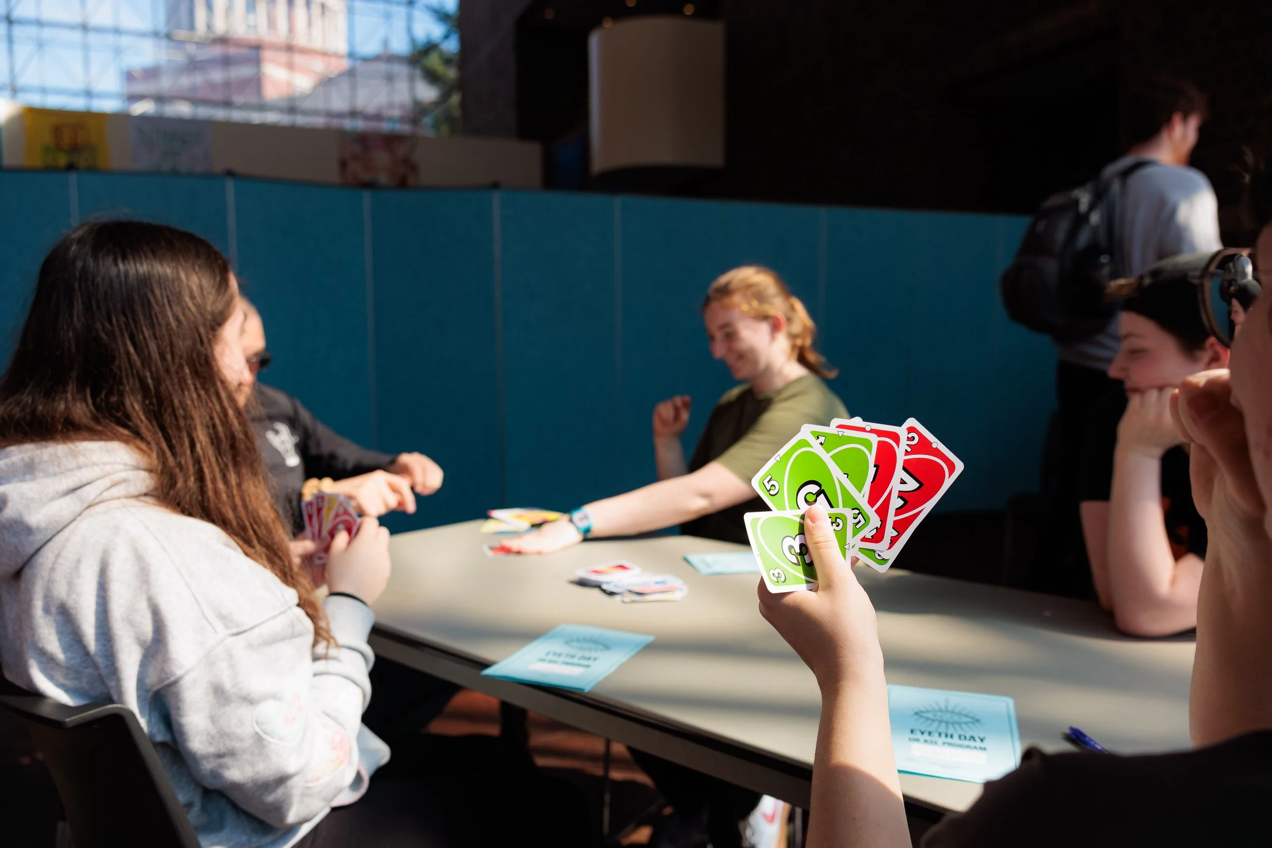 People playing Uno card game at a table, with one person holding a hand of colorful UNO cards. The setting appears to be indoors near a large window.