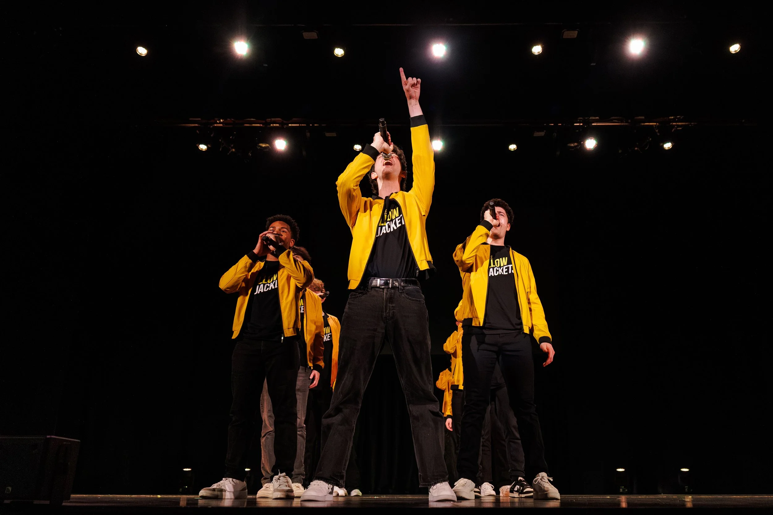 Group of performers on stage wearing yellow jackets and black t-shirts, with one person holding a microphone and raising their hand, performing under bright stage lights.
